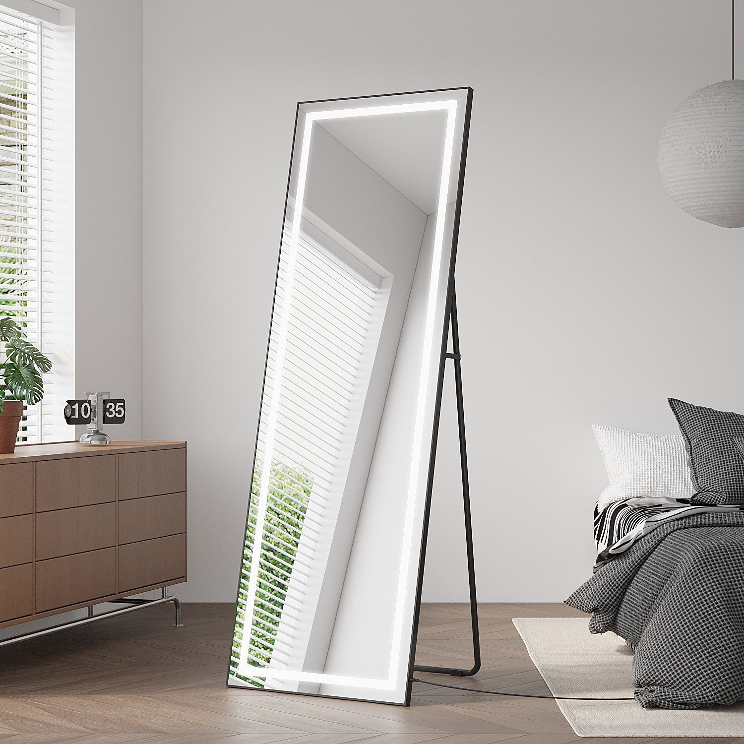 Ein hoher Spiegel mit leuchtendem Rahmen steht in einem Schlafzimmer. Eine Kommode, ein Bett und ein Fenster spiegeln sich.