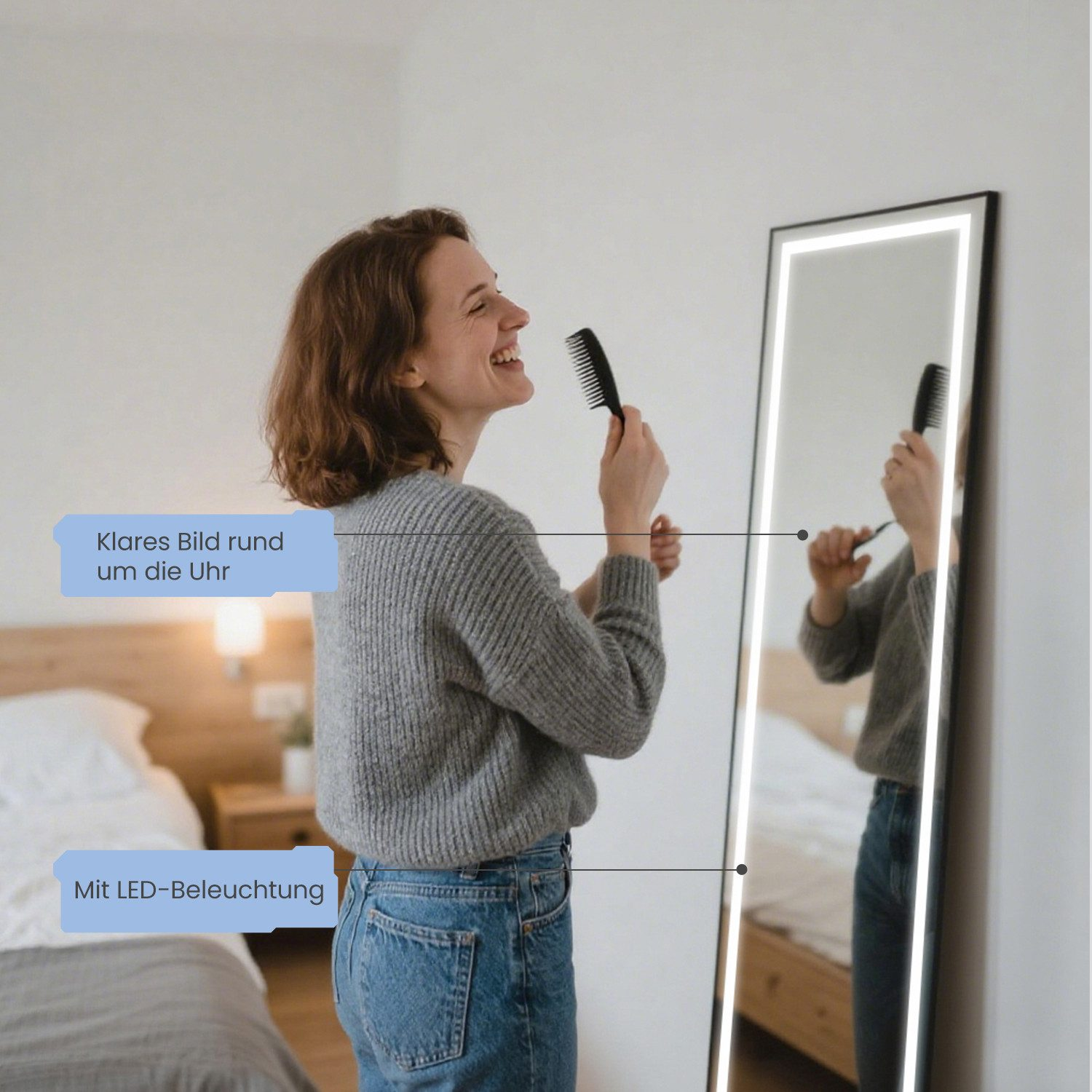 Eine Frau in grauem Pullover und Jeans kämmt sich vor einem beleuchteten Spiegel die Haare. Schlafzimmer mit Bett im Hintergrund.
