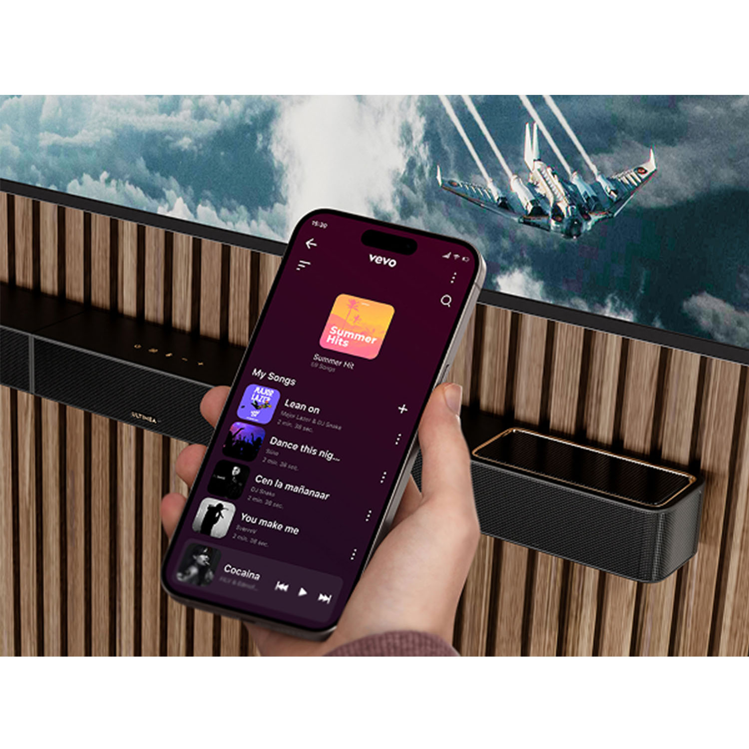 Ręka trzyma telefon z otwartą aplikacją muzyczną obok głośnika. Tło przedstawia samolot i drewnianą ścianę.