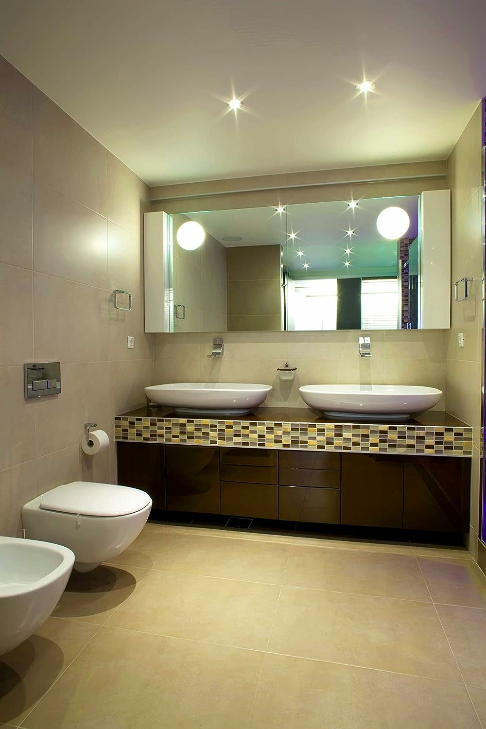 Baño moderno con dos lavabos, espejo grande y inodoro. Azulejos beige en paredes y suelo. Armarios marrones.