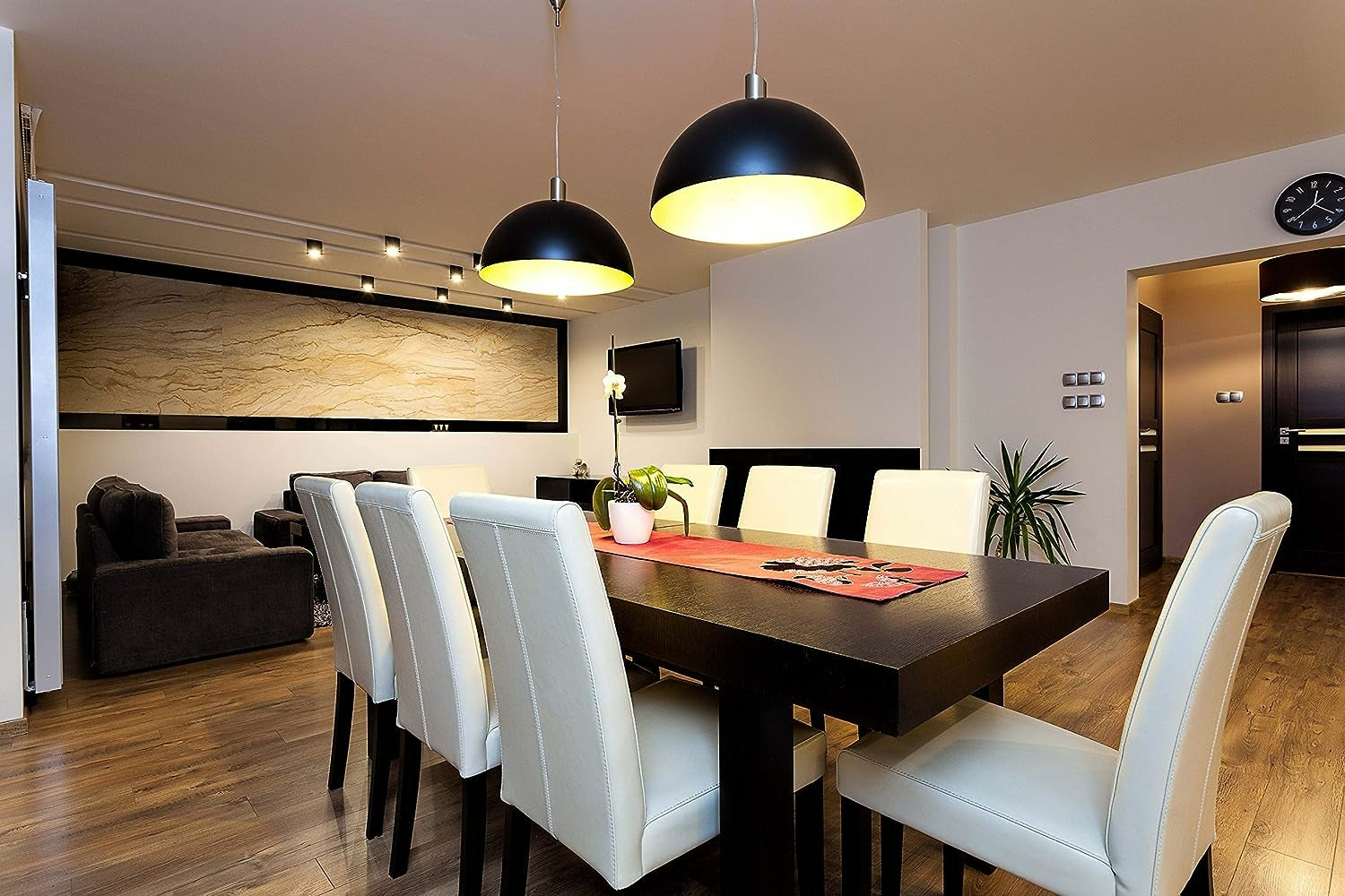 Un comedor moderno con mesa oscura, sillas blancas y lámparas colgantes negras. Un sofá en el fondo.
