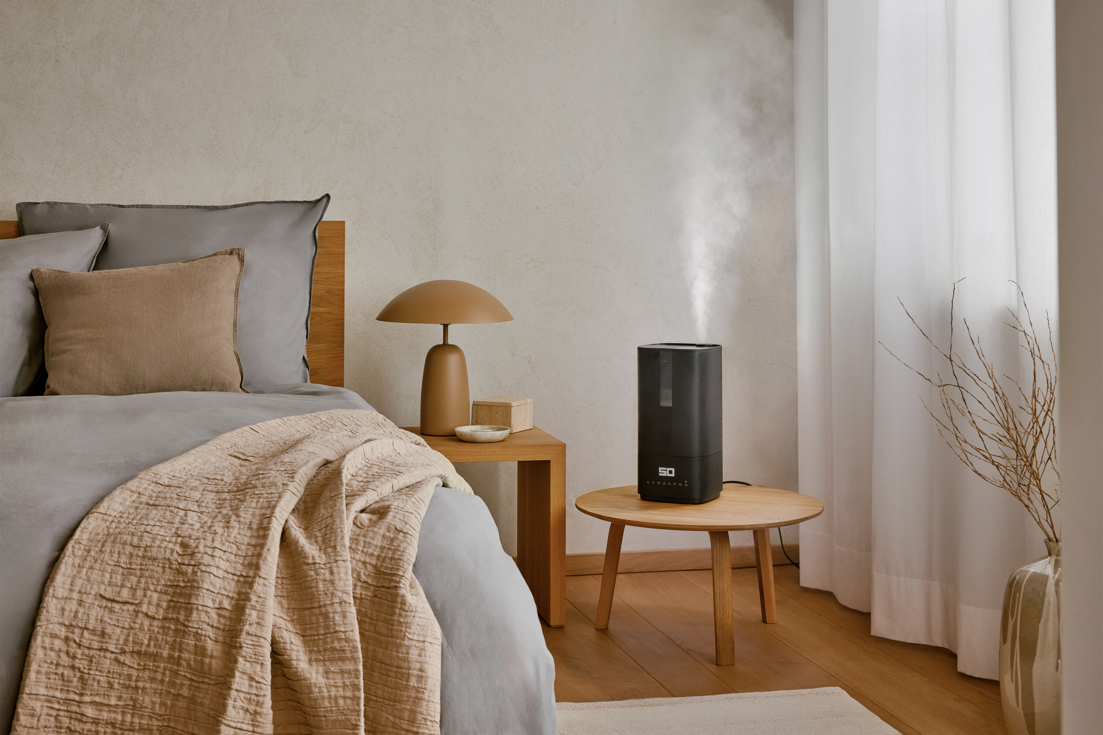 Bedroom scene with a bed, side tables, lamp, humidifier, and a vase with branches next to the window.