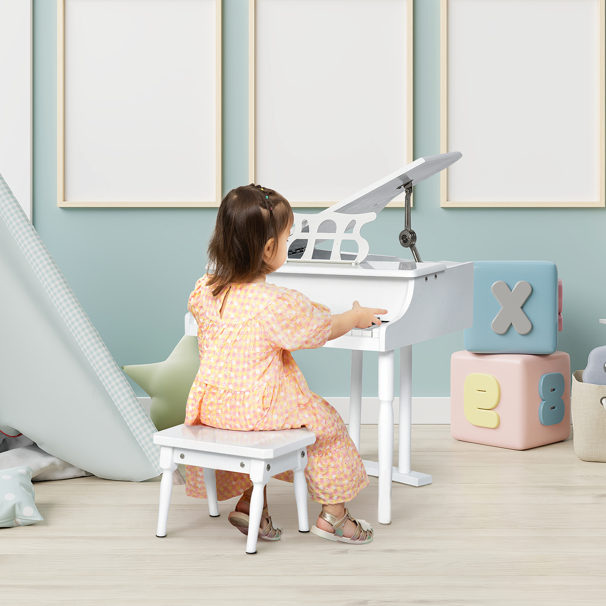 Una niña toca un piano de juguete blanco. Lleva un vestido floral y está sentada en un taburete pequeño. Se ven cubos y marcos en blanco.