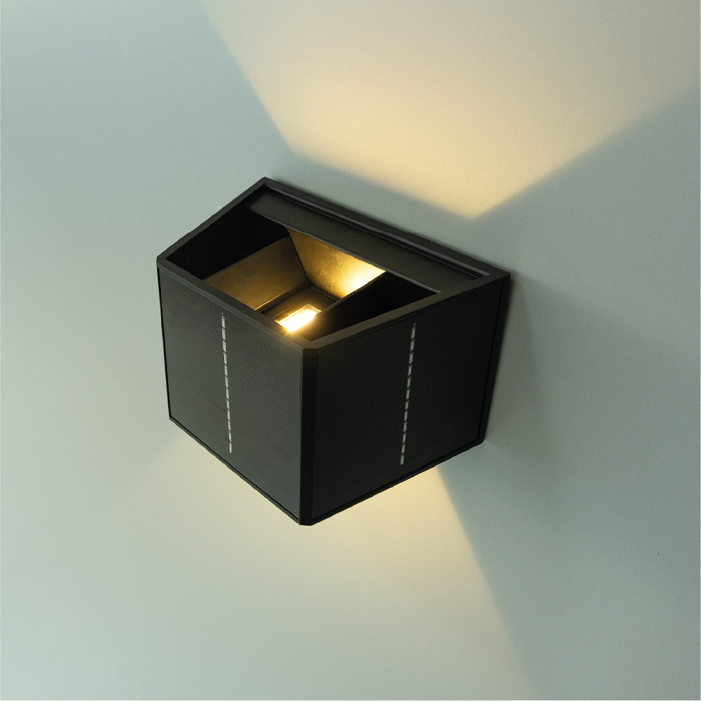 Una lámpara de pared negra y angular está montada en una pared gris claro. La lámpara emite luz cálida, proyectando sombras. La lámpara tiene una superficie texturizada.