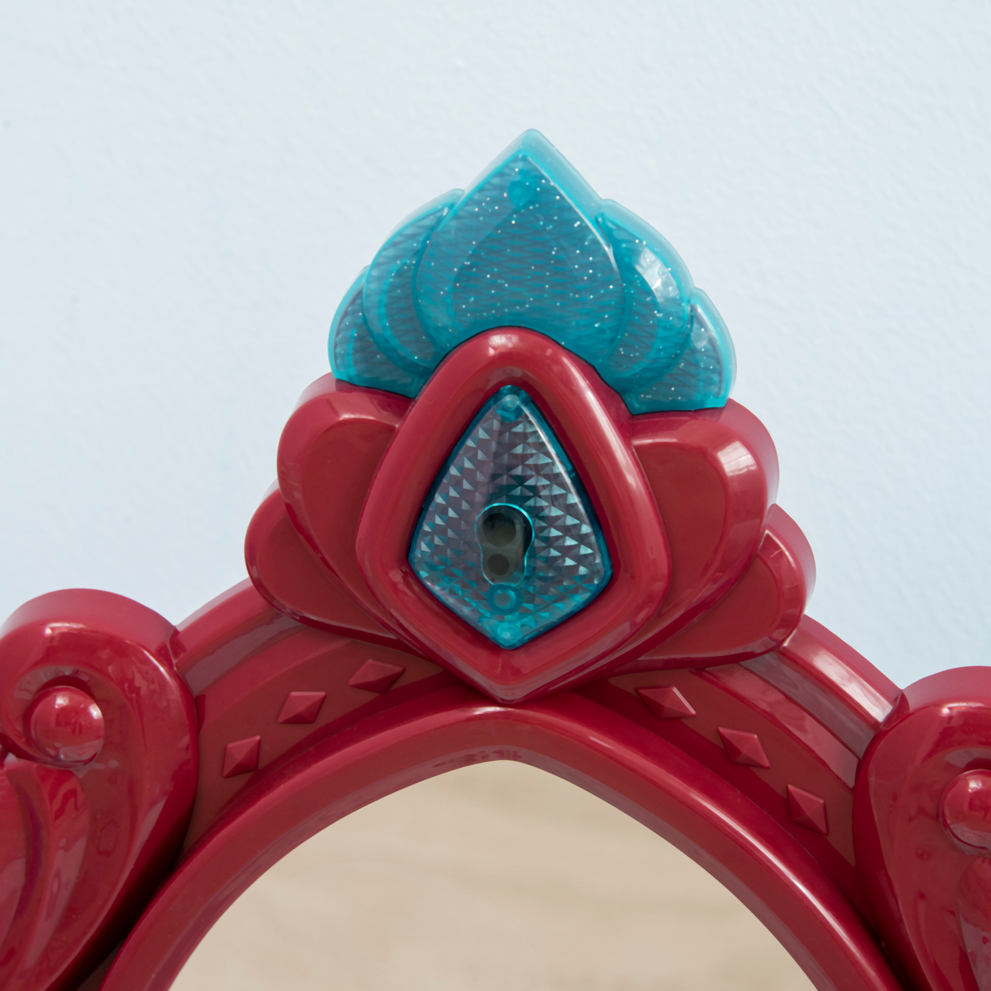 Un espejo de juguete de plástico rojo y azul. El espejo tiene forma de ojo de cerradura y una gema azul. El fondo es azul claro.