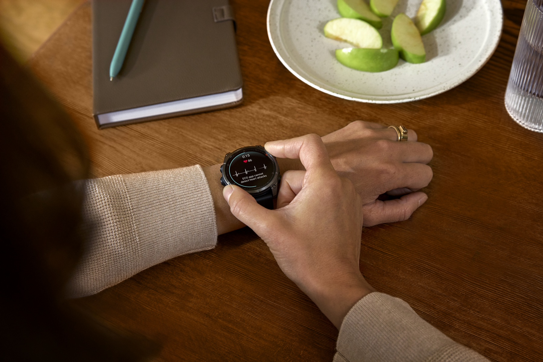 Poignet avec montre connectée affichant la fréquence cardiaque. Tranches de pommes sur une assiette, un carnet et un stylo visibles.