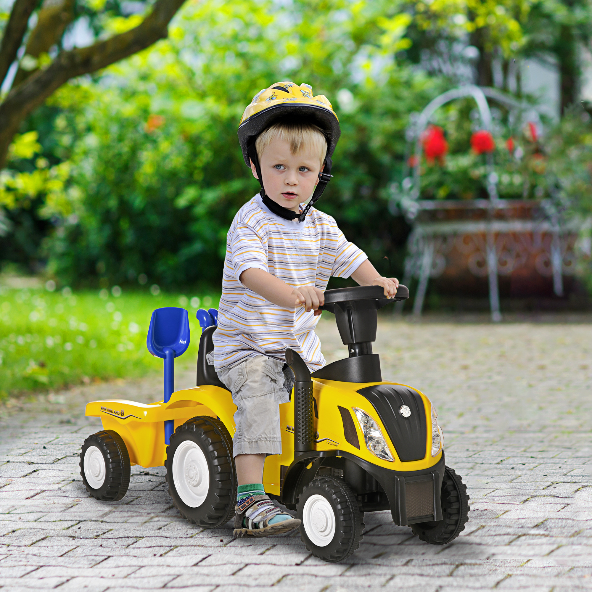 Un niño pequeño con casco está sentado en un tractor de juguete amarillo. El tractor está en un patio de ladrillos, con vegetación al fondo.