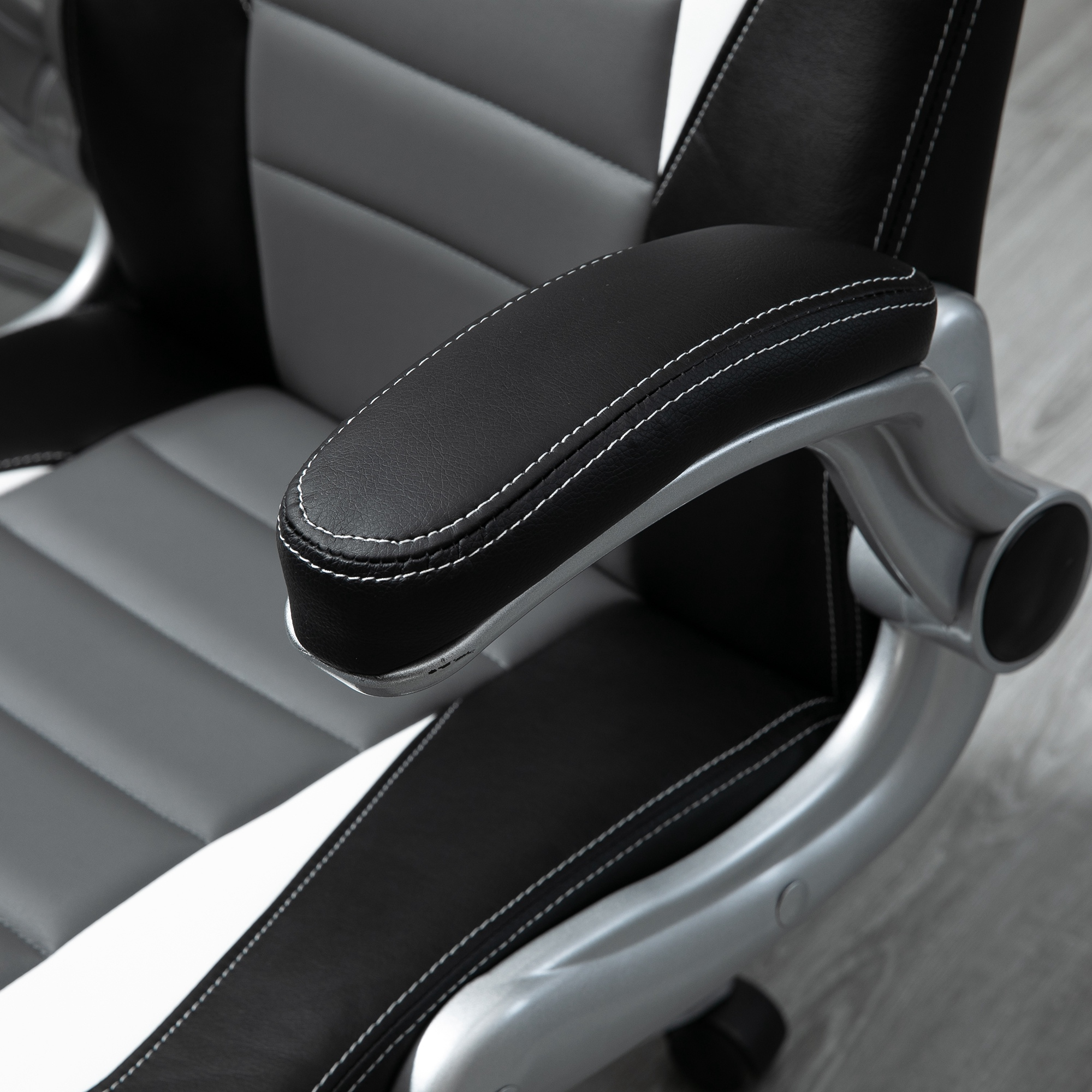 Primer plano del reposabrazos de una silla gaming. Cuero negro con costuras blancas y detalles grises. El fondo es un suelo de madera.