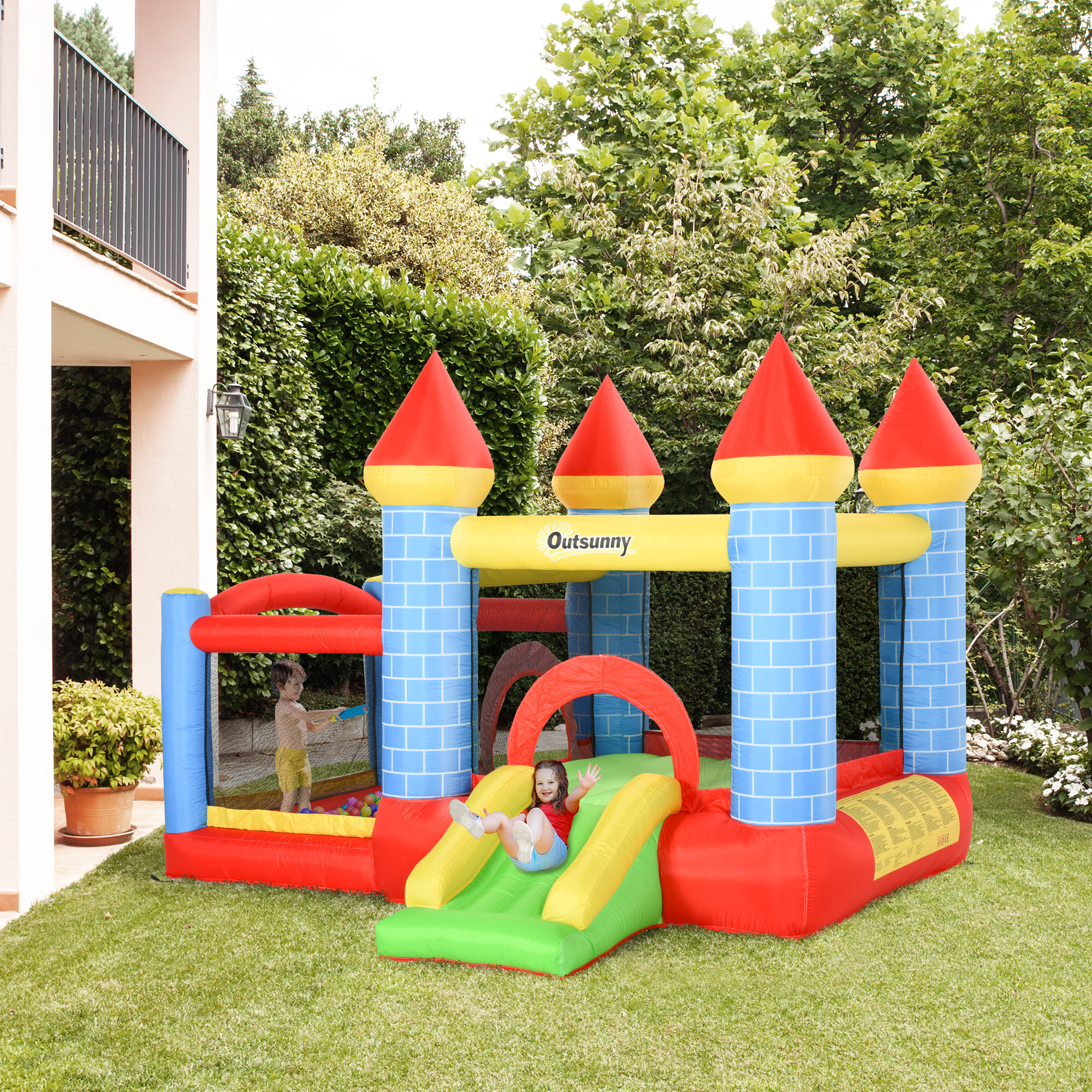 Un castillo hinchable con colores rojo, azul y amarillo está sobre una hierba verde. Dos niños están jugando. Hay árboles y una casa detrás.