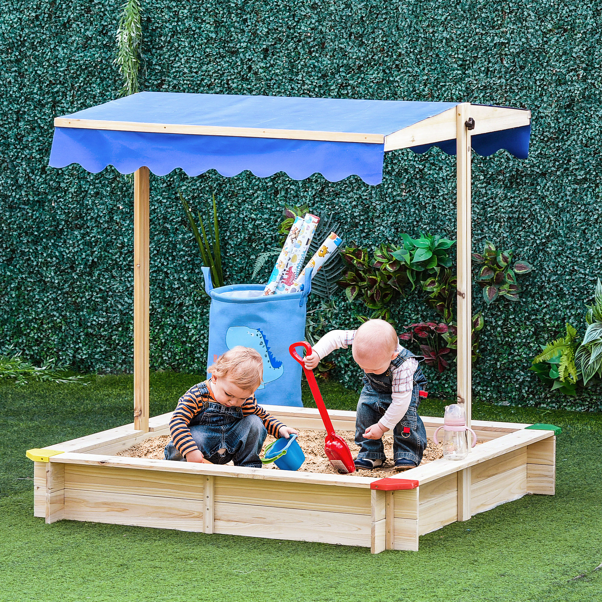 Dos niños pequeños juegan en un arenero bajo un toldo. Uno usa una pala, el otro un cubo. Fondo de seto verde.