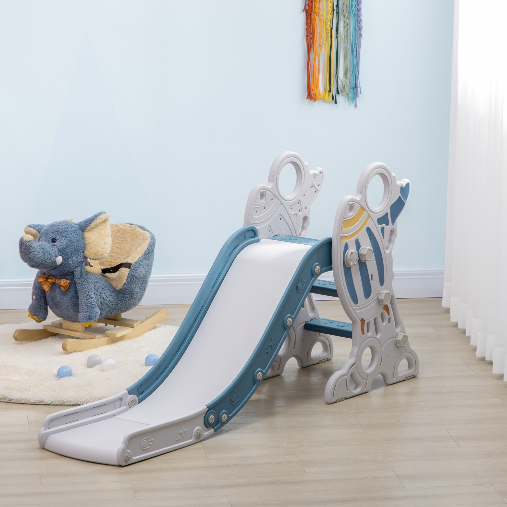 Un tobogán de plástico azul y blanco. Se ven un balancín de elefante y una colorida decoración de pared. El suelo es de madera clara.