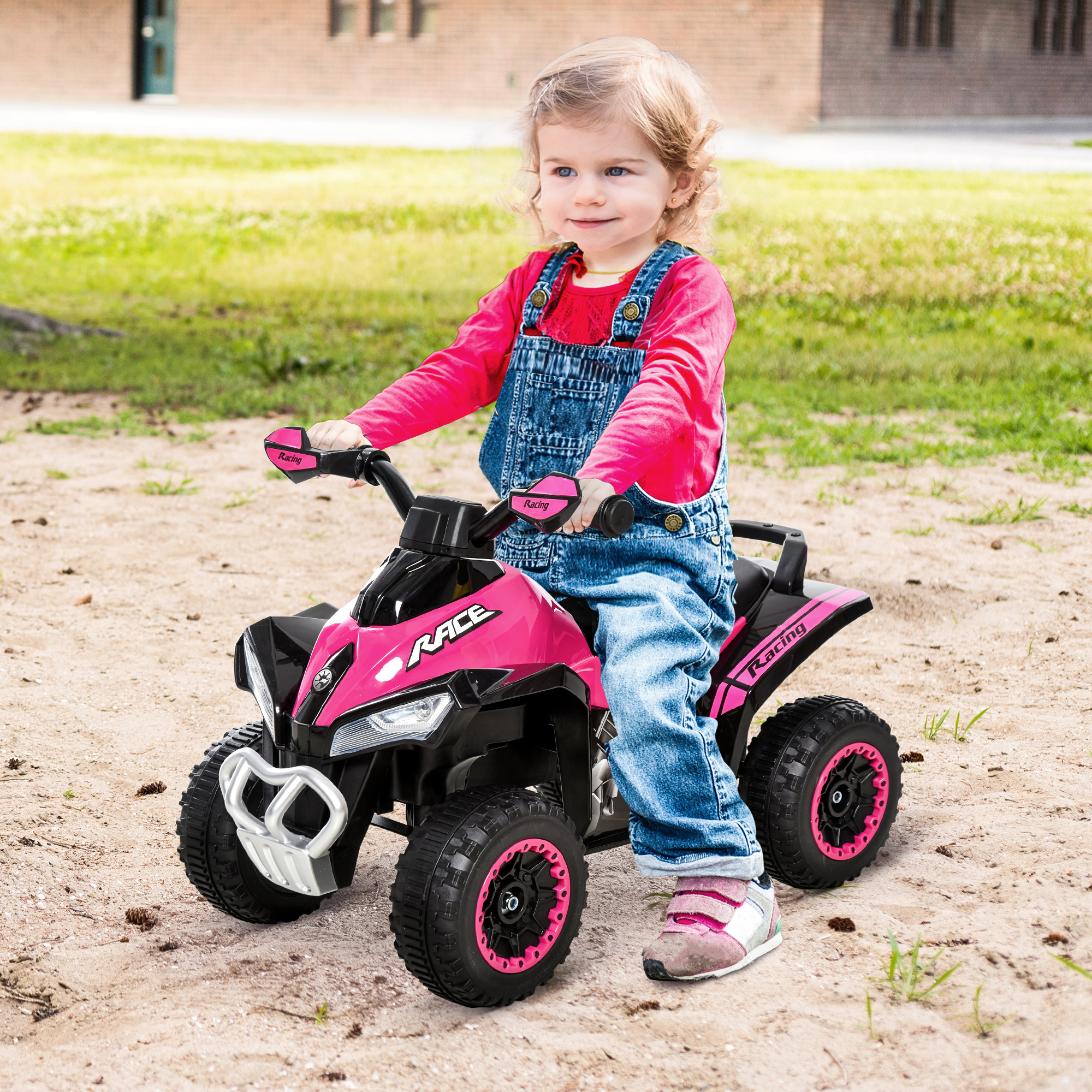 Un niño con overoles monta un quad de juguete rosa y negro sobre arena. Un edificio de ladrillo y hierba en el fondo.
