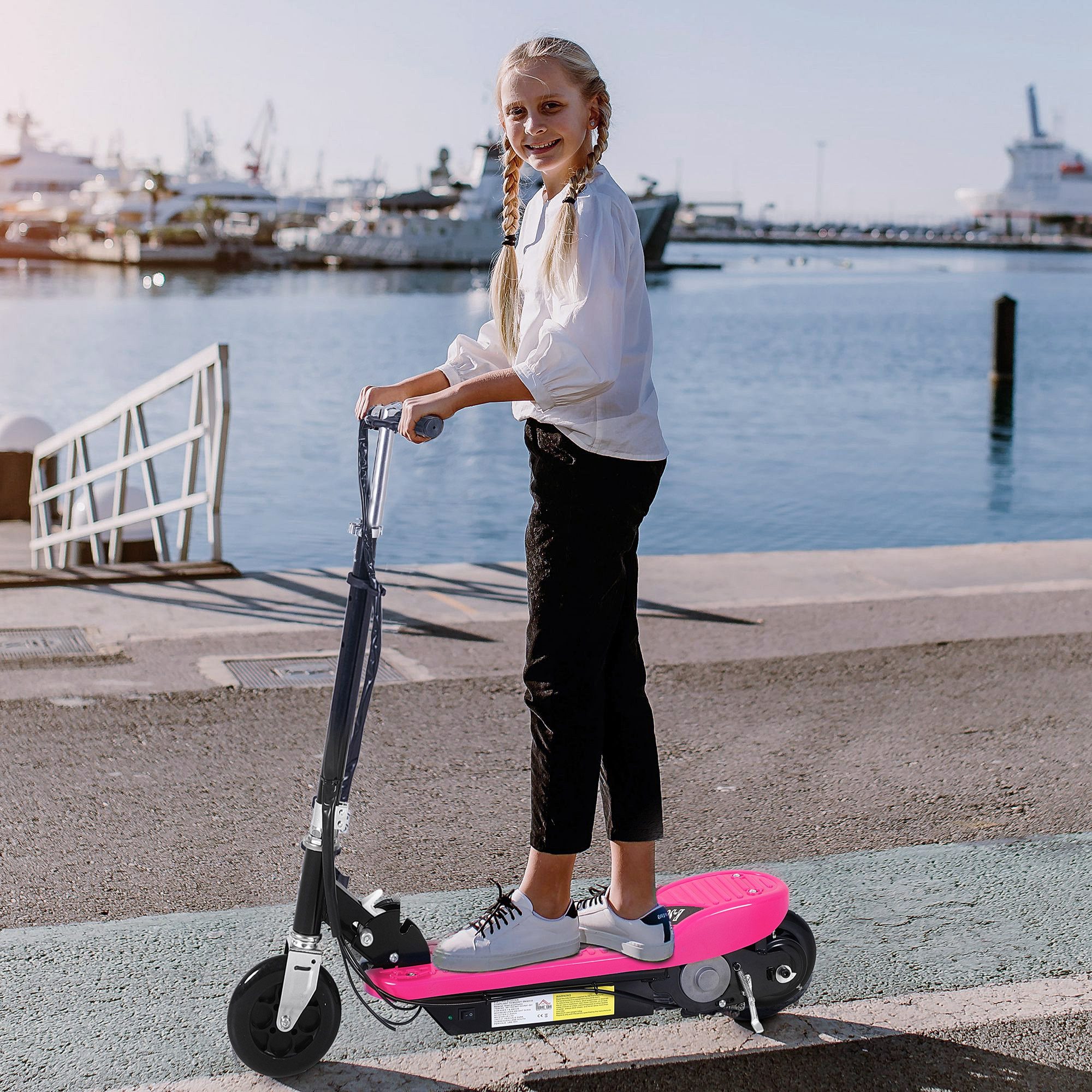Una niña en un scooter rosa sonríe. Lleva camisa blanca y pantalón negro. El fondo es un mar azul y barcos.