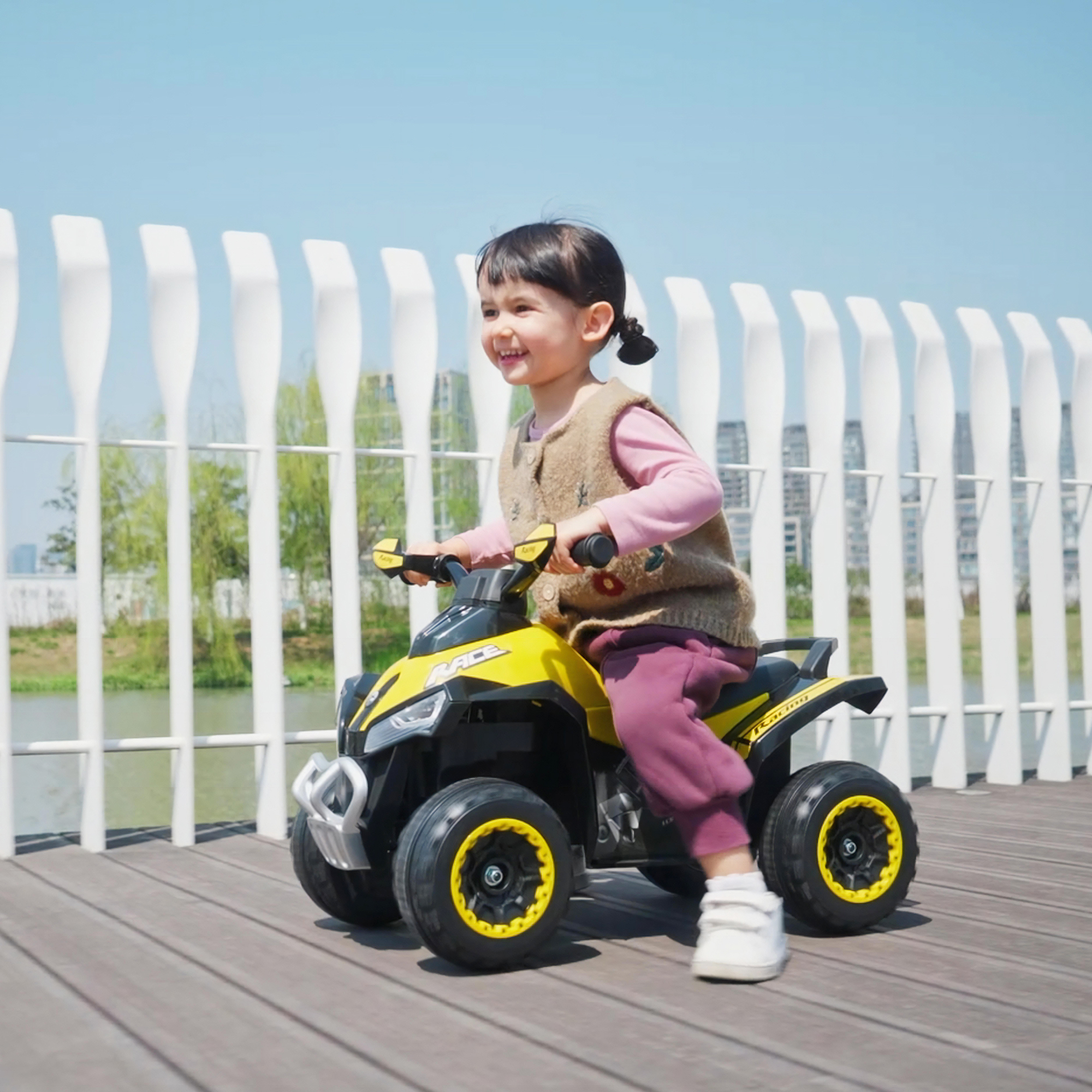 Una niña sonríe mientras monta un quad de juguete amarillo y negro en un camino de madera. Valla blanca y cielo azul al fondo.