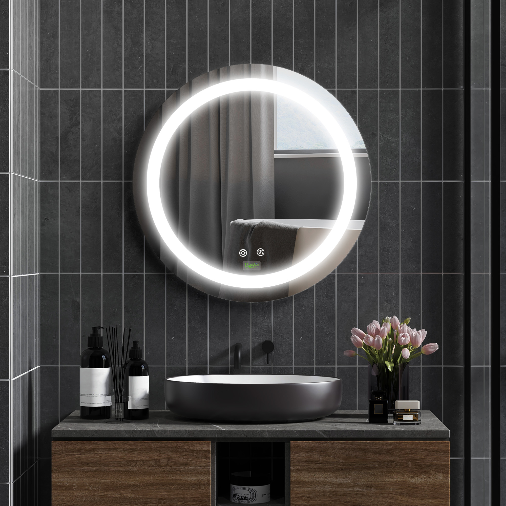 Un baño con espejo redondo, lavabo negro y mueble de madera. Flores y artículos de tocador están en la encimera.