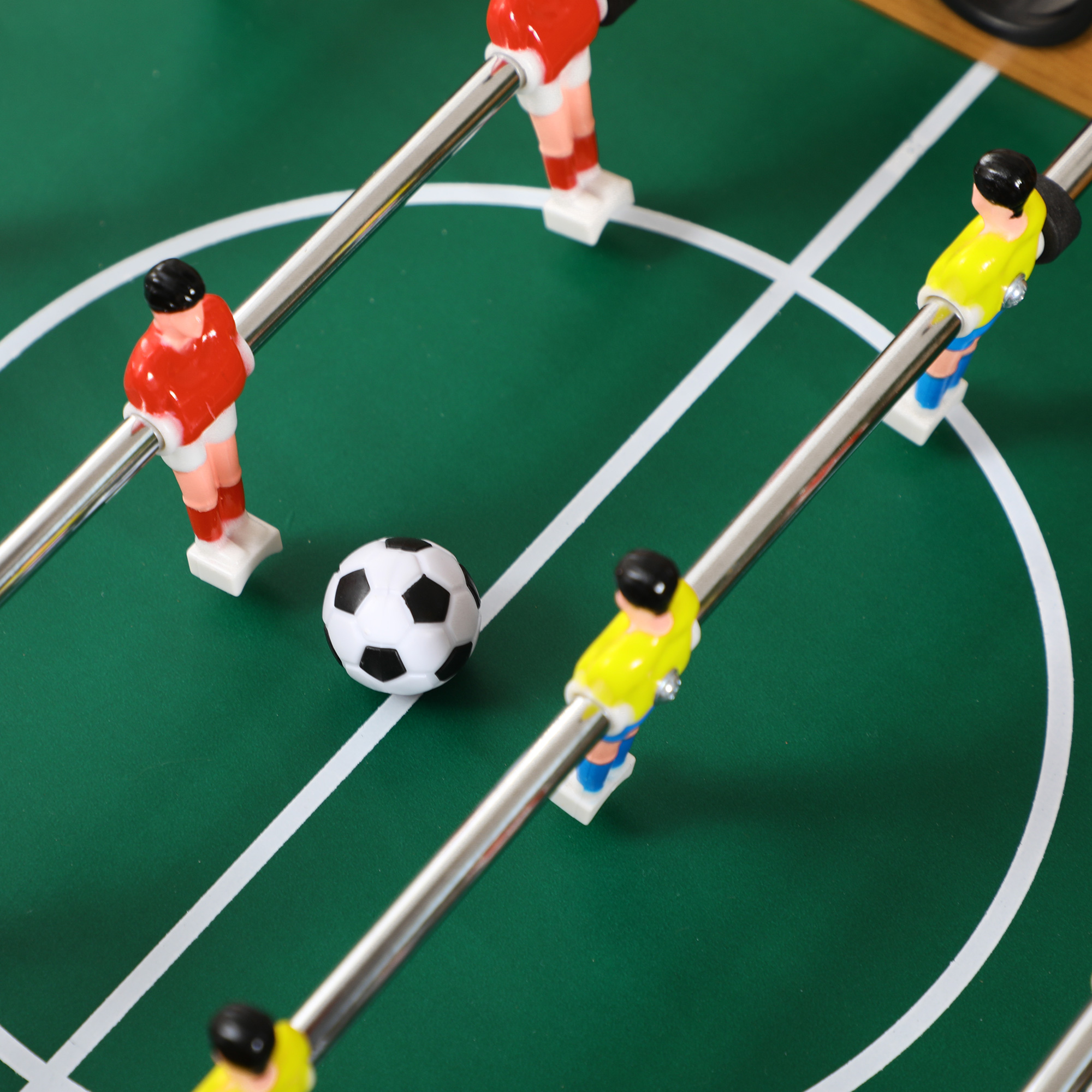 Primer plano de futbolín: jugadores rojos y amarillos, balón blanco y negro, sobre campo verde con líneas blancas.