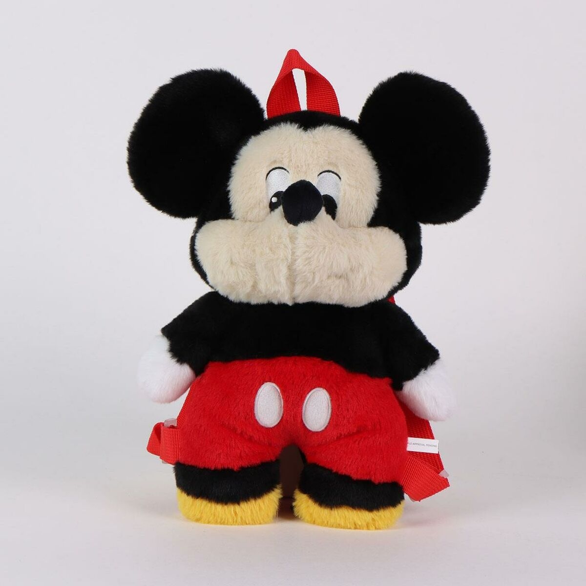 Una mochila de peluche de Mickey Mouse. Mickey tiene orejas negras, cara blanca, pantalones cortos rojos y una correa roja. El fondo es blanco.