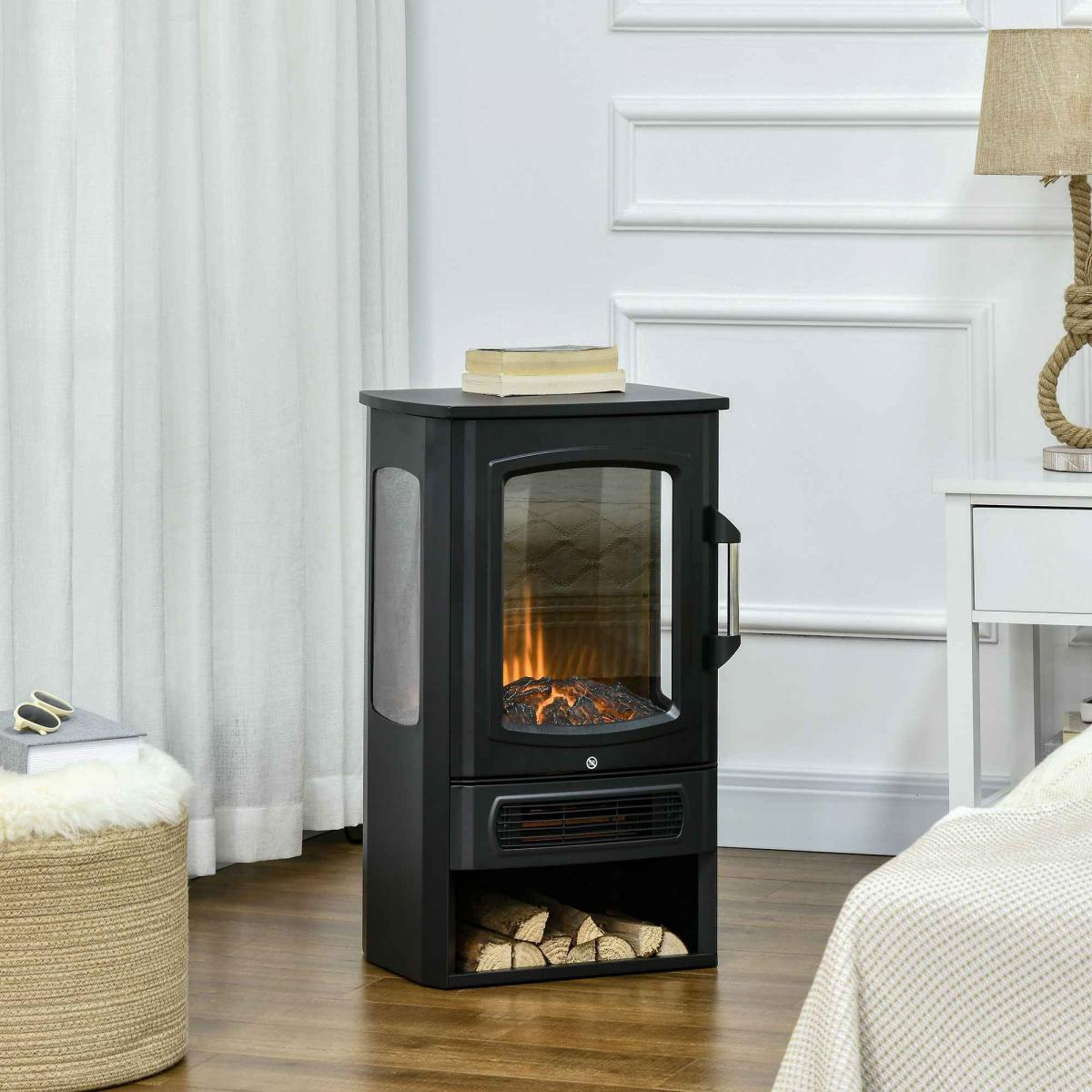 Schwarzer Elektro-Kamin mit Glasfront und Holzscheiten. Daneben ein weißer Tisch mit Lampe. Hocker und Bett sichtbar.
