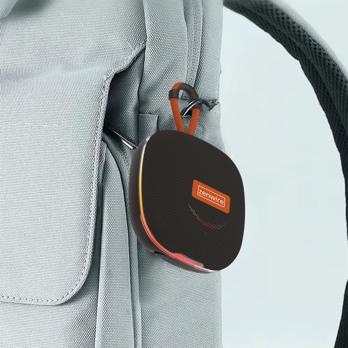 Czarny głośnik z pomarańczowym logo jest przymocowany do szarego plecaka za pomocą karabińczyka. Głośnik ma kolorowe światła.