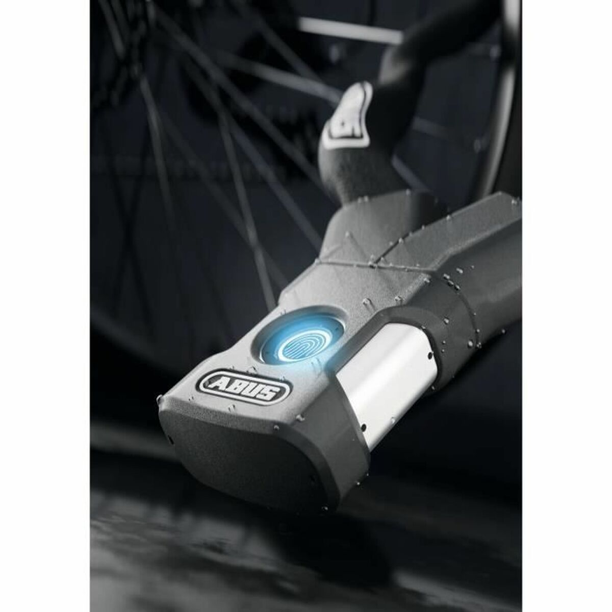 Nahaufnahme eines schwarzen und grauen Fahrradschlosses mit blau leuchtendem Fingerabdruckscanner, ABUS-Logo sichtbar. Fahrradrad im Hintergrund.
