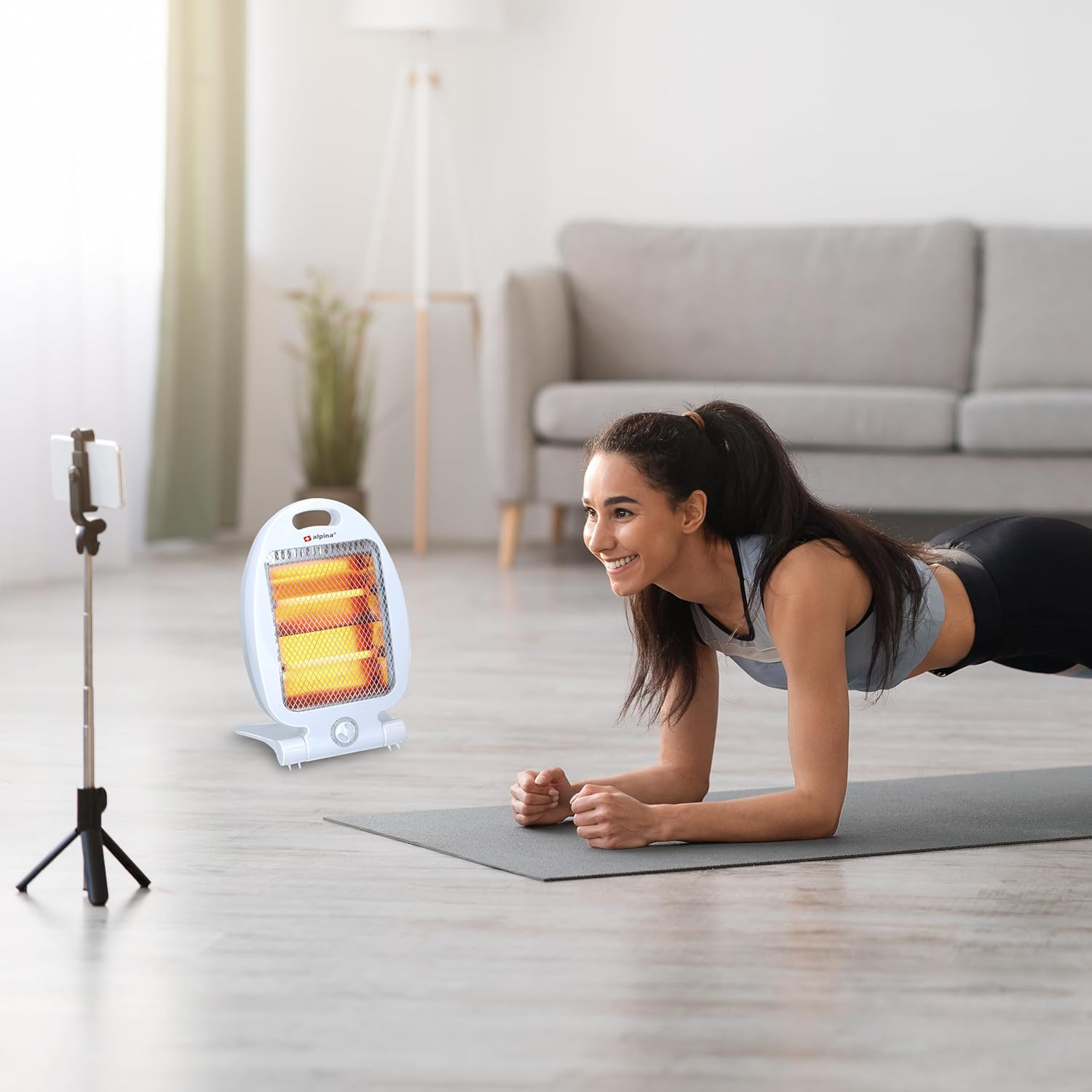 Eine Frau in der Plank-Pose auf einer Matte, lächelnd. Ein Heizgerät und ein Telefon auf einem Stativ sind im Hintergrund.
