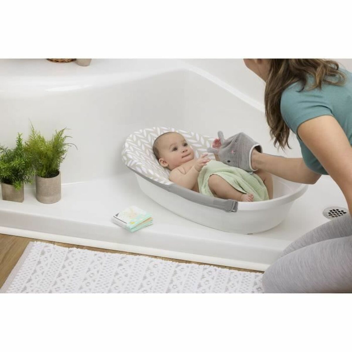 Ein Baby liegt in einer weißen Wanne mit einer gemusterten Einlage. Eine Frau wäscht das Baby mit einem Waschlappen. Pflanzen, ein Buch und ein Teppich sind sichtbar.