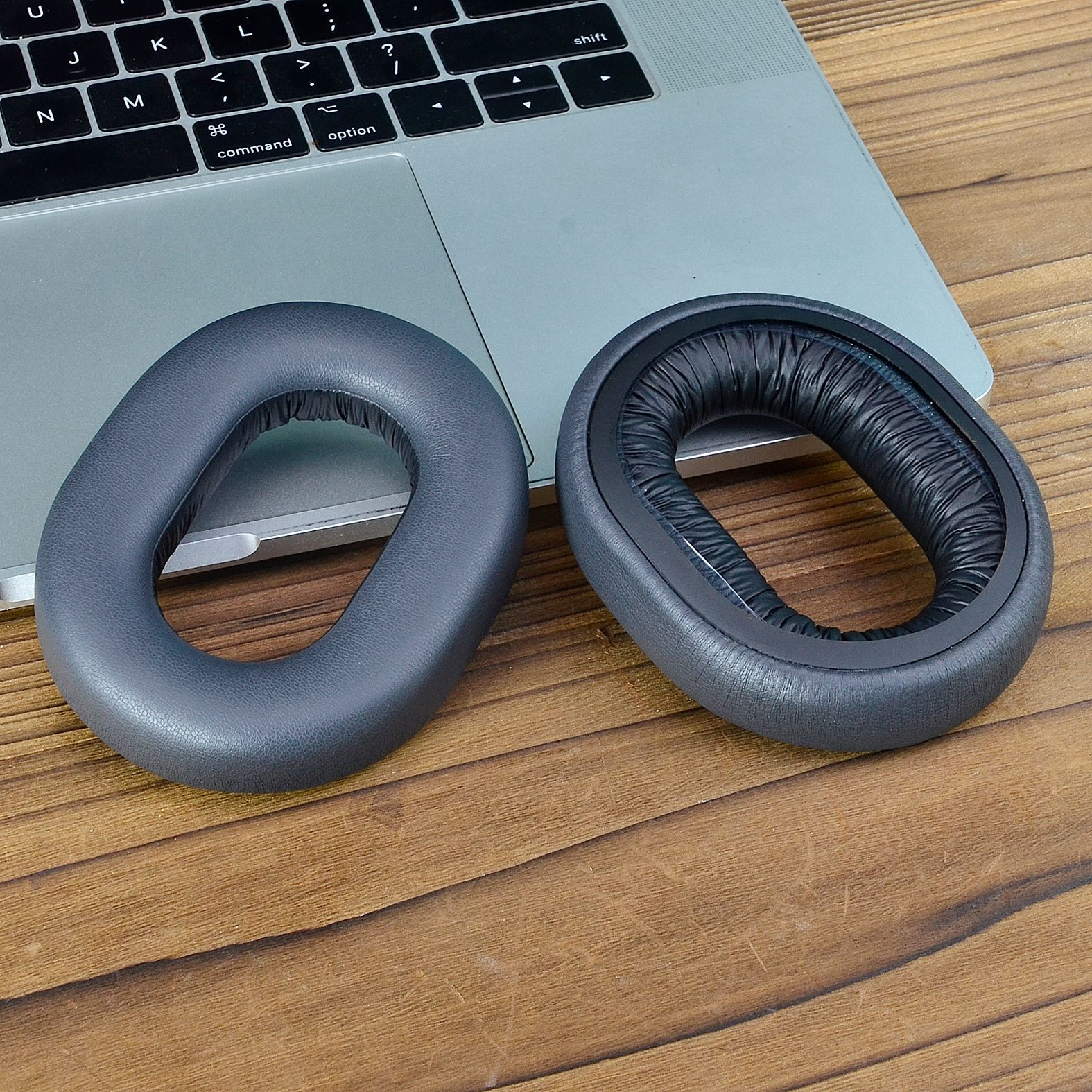 Dwie szare poduszki na uszy do słuchawek na drewnianym stole, z laptopem w tle.