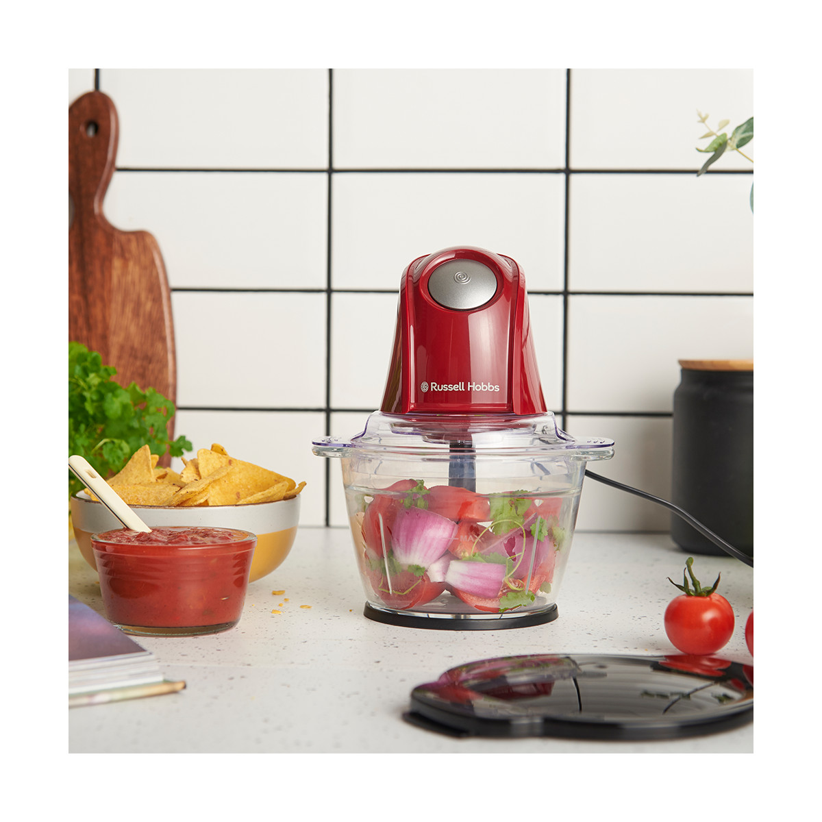 Ein roter Foodprocessor mit Gemüse, Salsa, Chips und einer Tomate auf einer weißen Arbeitsplatte. Im Hintergrund ein Schneidebrett.