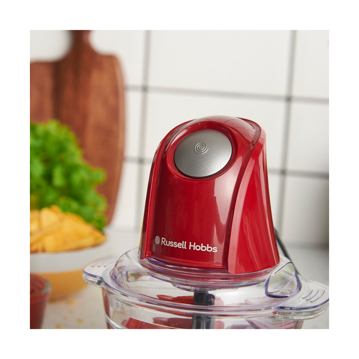 Ein roter Russell Hobbs Zerkleinerer. Er hat einen silbernen Knopf oben. Im Hintergrund sind eine Schüssel mit Essen und ein Holzbrett.