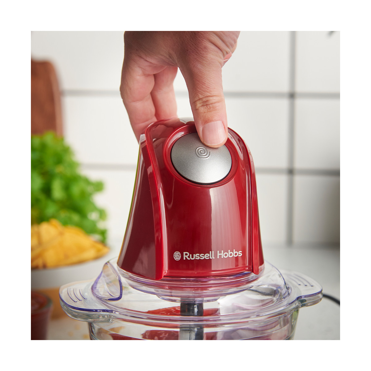 Eine Hand drückt auf die Taste eines roten Russell Hobbs Zerkleinerers. Der Zerkleinerer steht auf einer Glasschüssel mit Essen. Im Hintergrund Essen und Fliesen.