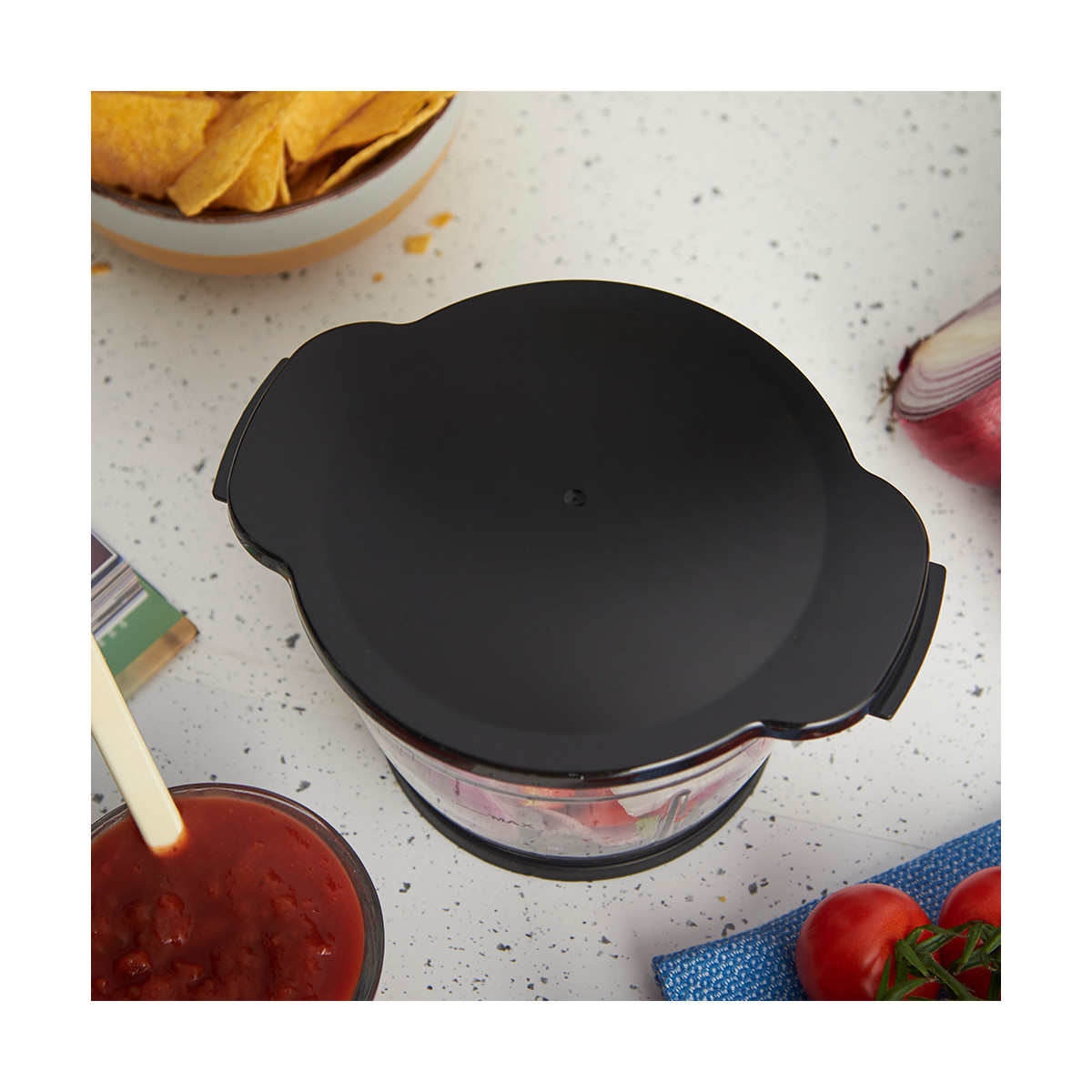 Ein Foodprocessor mit schwarzem Deckel, Salsa, Tomaten, Chips und roter Zwiebel auf einer gesprenkelten Theke.