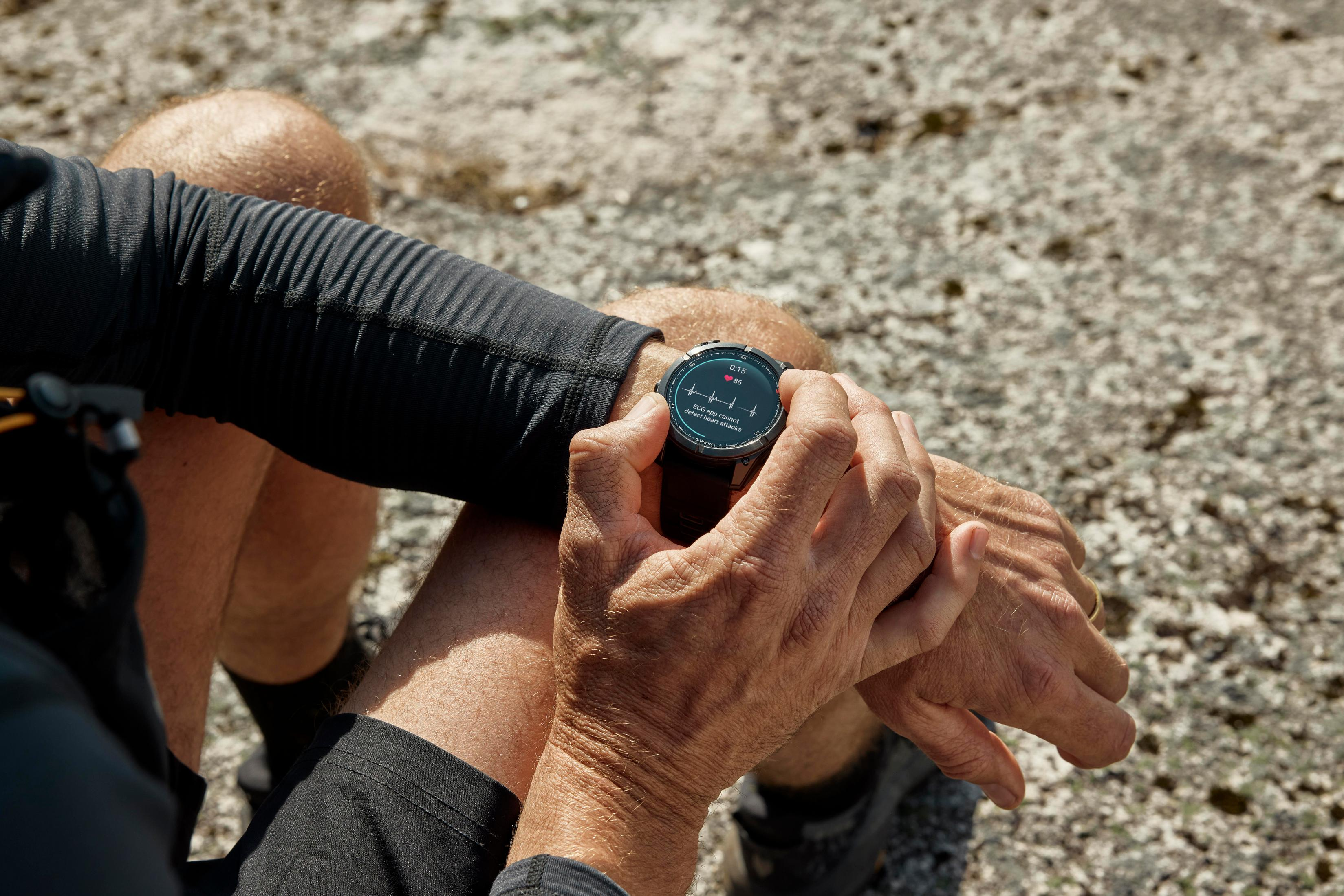 Handgelenk mit Smartwatch, die Zeit und Herzfrequenz anzeigt. Der Hintergrund sind verschwommene Felsen.