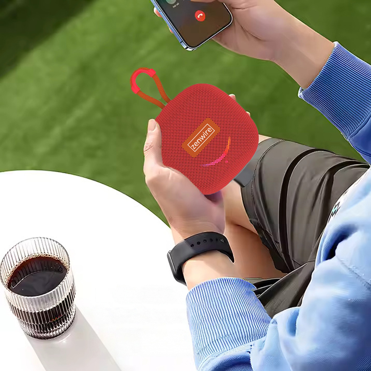 Osoba trzymająca czerwony głośnik Zenwire i telefon. Szklanka kawy na stole, z trawą w tle.