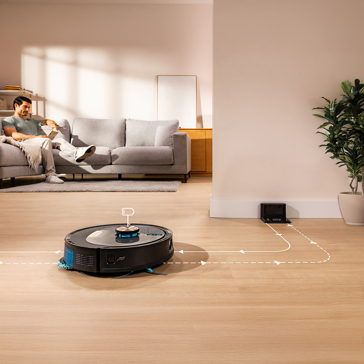 Un uomo si rilassa su un divano mentre un aspirapolvere robot pulisce il pavimento. Una stazione di ricarica è visibile sullo sfondo.