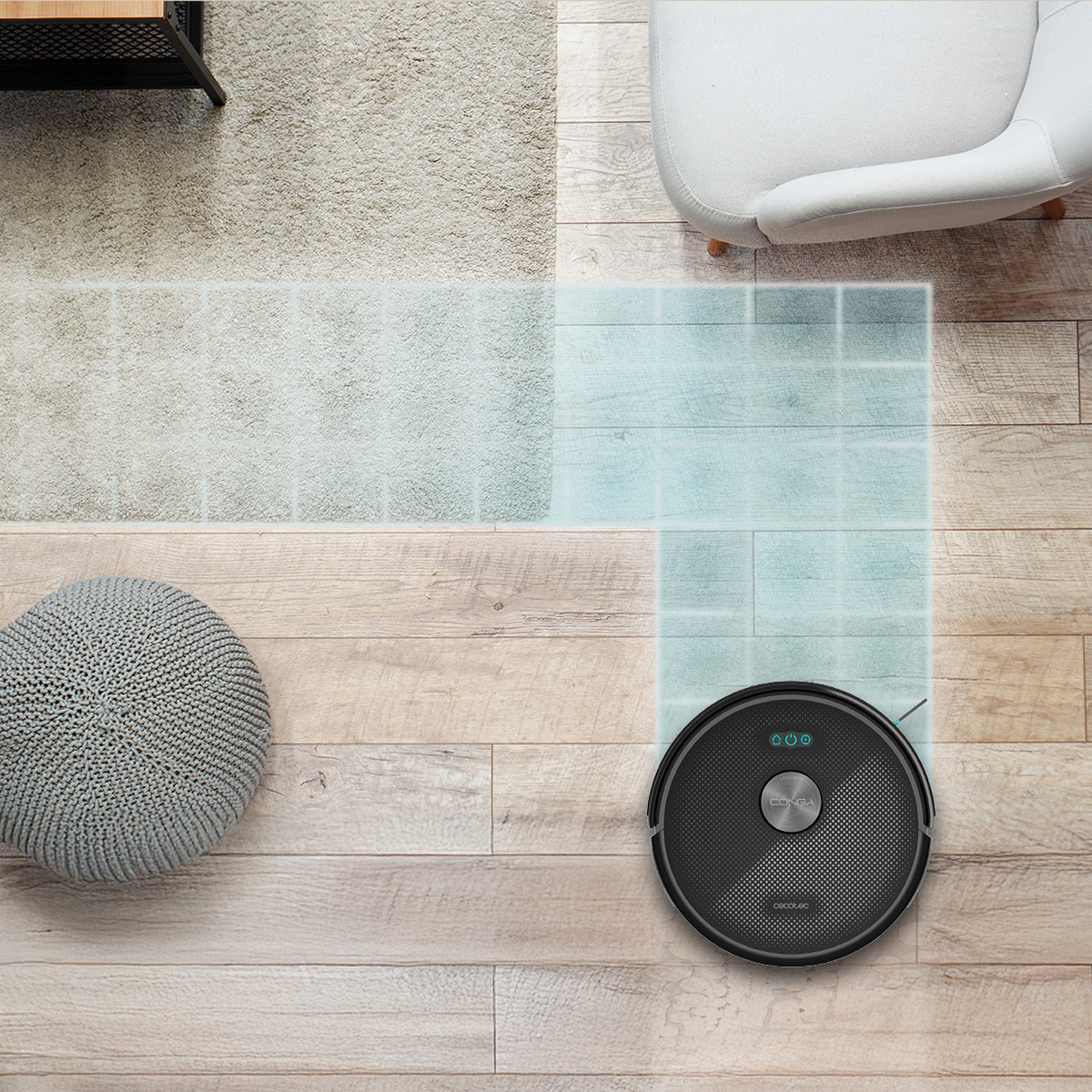 Un aspirapolvere robot nero su pavimento in legno. Sono visibili anche un pouf grigio, una poltrona bianca e un tappeto beige.
