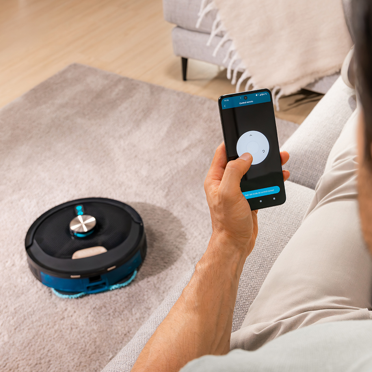 Una persona tiene in mano uno smartphone che controlla un robot aspirapolvere. L'aspirapolvere è sul tappeto. Un divano è sullo sfondo.