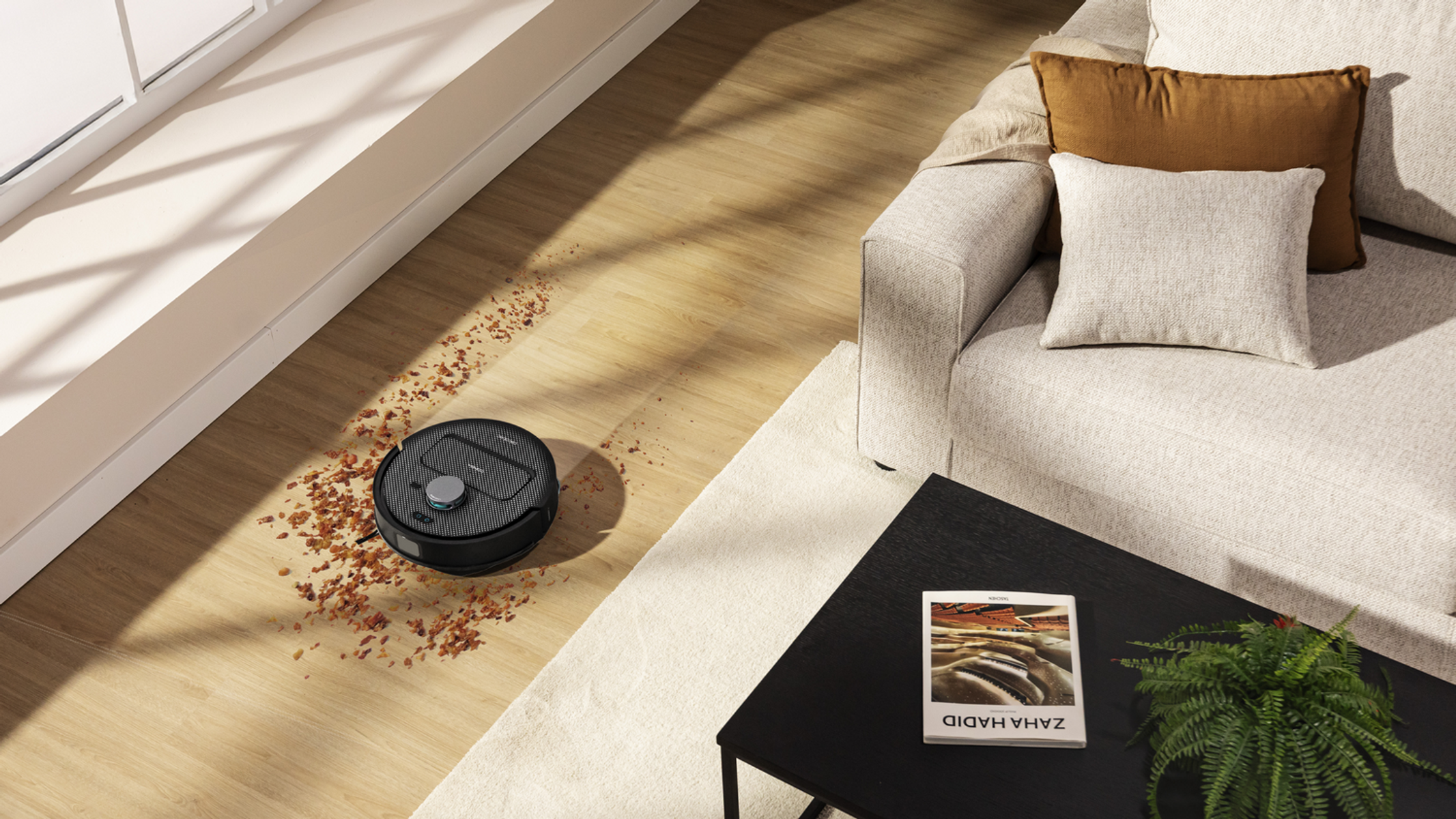 Un robot aspirapolvere pulisce un pavimento in legno con detriti. Sullo sfondo ci sono un divano, un tavolino e una finestra.