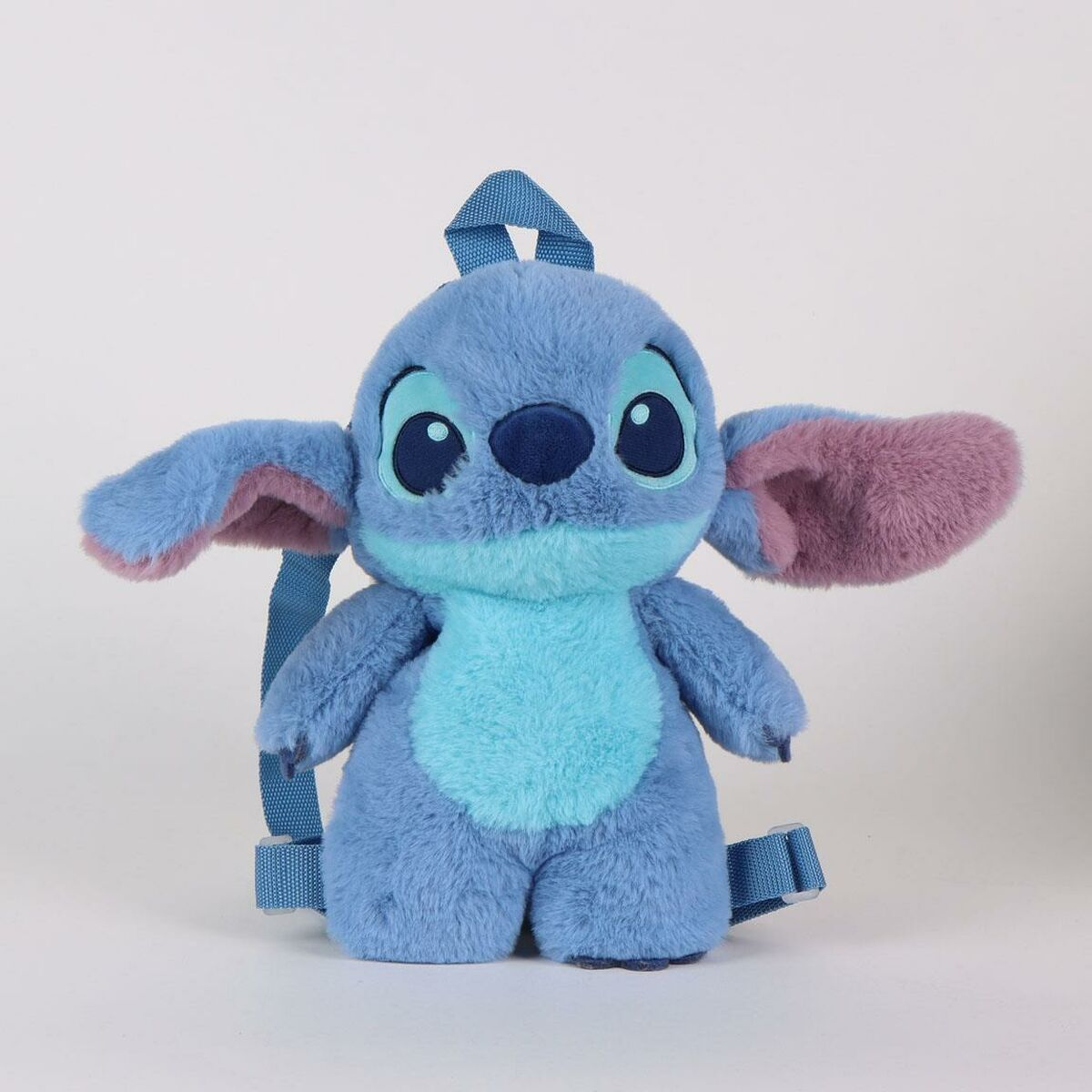 Una mochila de peluche azul de Stitch está sobre una superficie blanca. Tiene orejas grandes con interior rosa y correas azules.