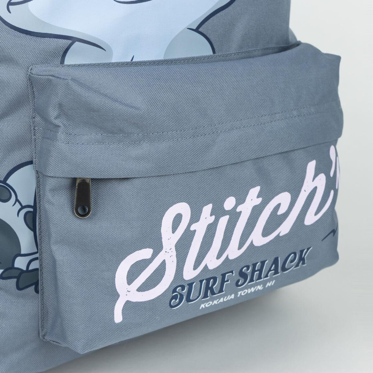 Una mochila gris con un bolsillo frontal. Presenta la palabra 'Stitch' y 'Surf Shack' en blanco, y un diseño de personaje de dibujos animados.