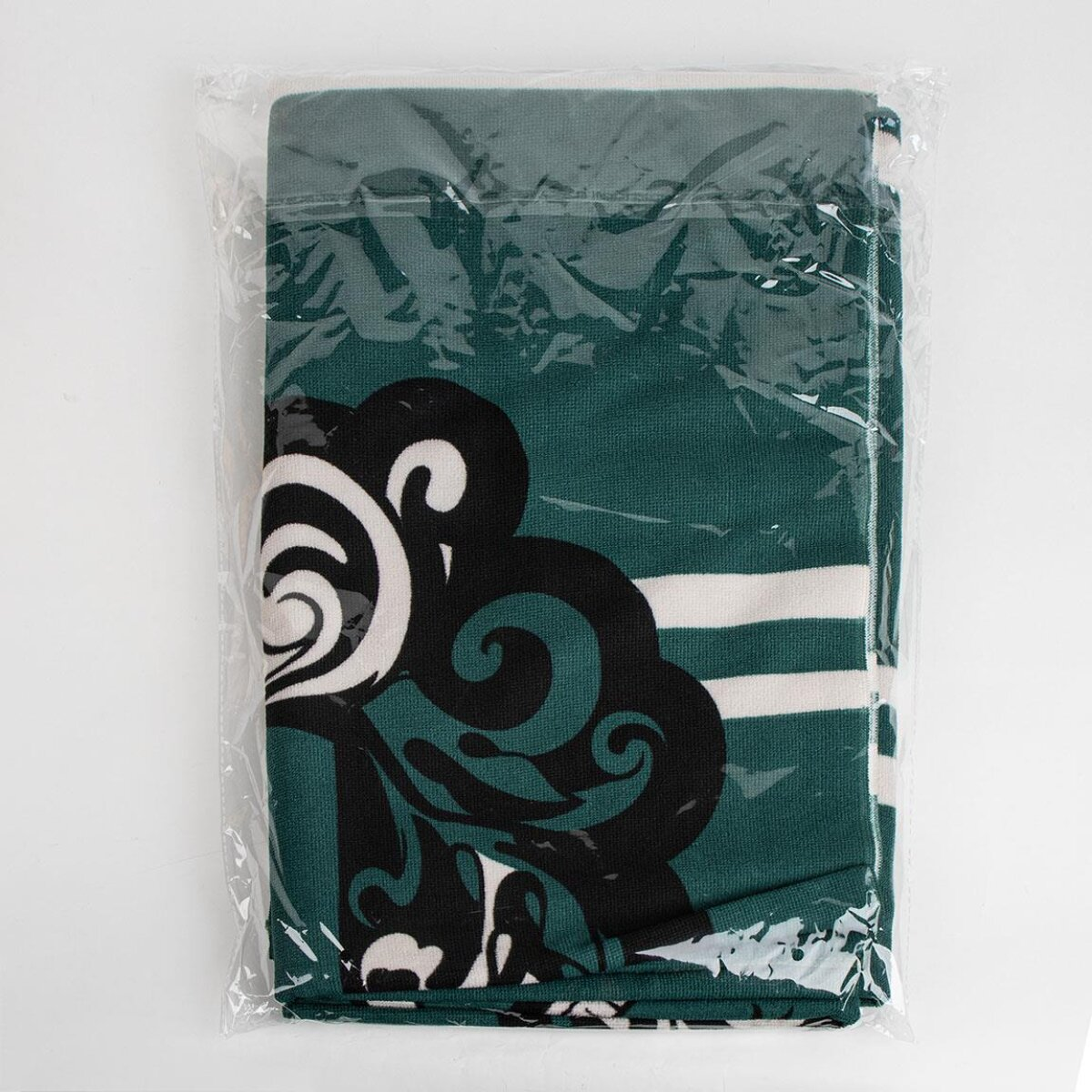 Una toalla verde doblada con diseños en blanco y negro está dentro de una bolsa de plástico transparente. Fondo blanco.