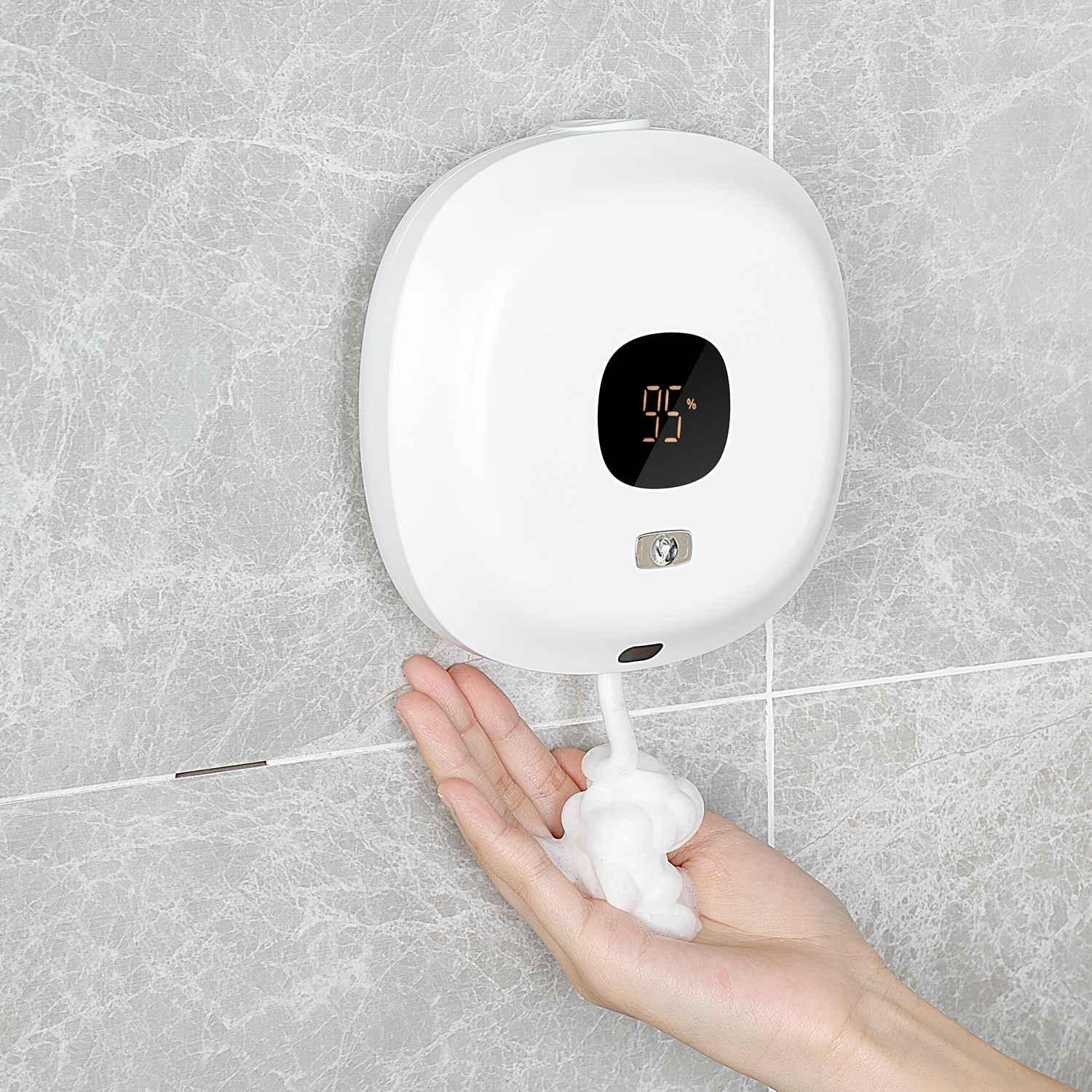 Ein weißer automatischer Seifenspender an einer grauen gefliesten Wand. Eine Hand empfängt Schaumseife. Der Spender zeigt 95% an.