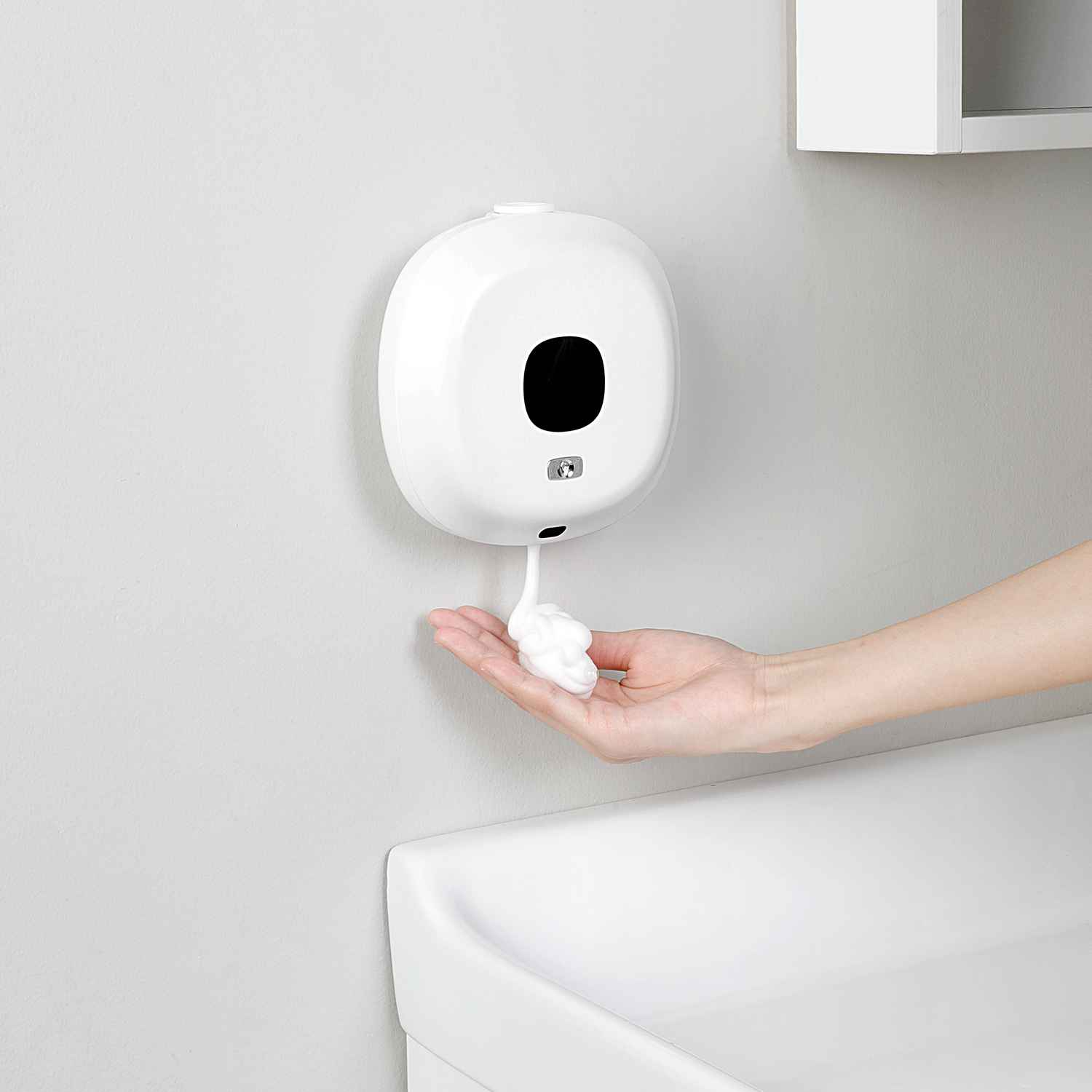 Ein weißer automatischer Seifenspender gibt Schaum in eine Hand ab. Der Spender ist an einer grauen Wand über einem weißen Waschbecken montiert.