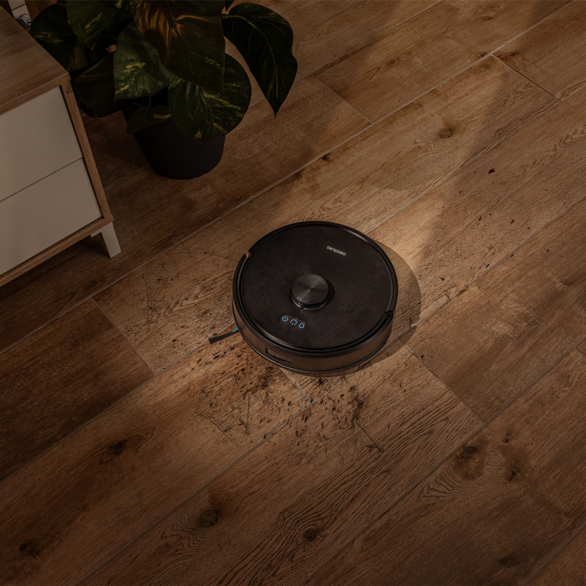 Un aspirapolvere robot nero su un pavimento in legno. Una pianta e un mobile sono sullo sfondo. Il pavimento è sporco.
