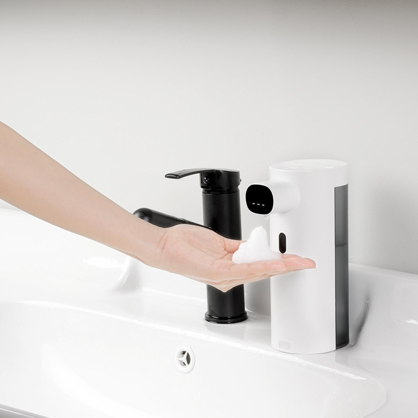 Eine Hand empfängt Schaum von einem automatischen Seifenspender in Weiß und Schwarz. Links ist ein schwarzer Wasserhahn.