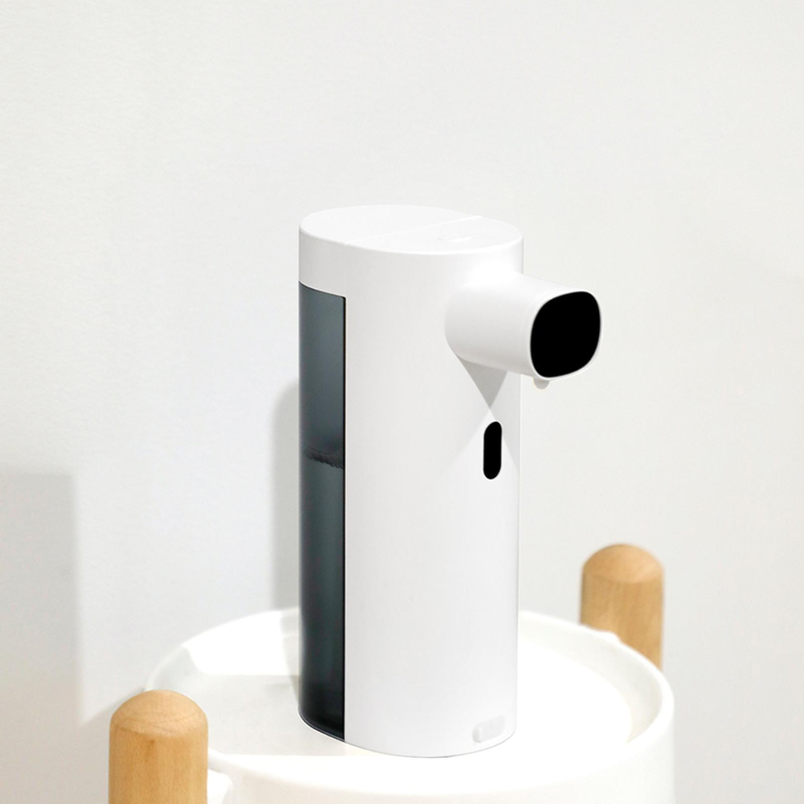 Weißer automatischer Seifenspender mit schwarzer Düse. Er steht auf einer weißen Oberfläche mit Holzbeinen. Weißer Hintergrund.