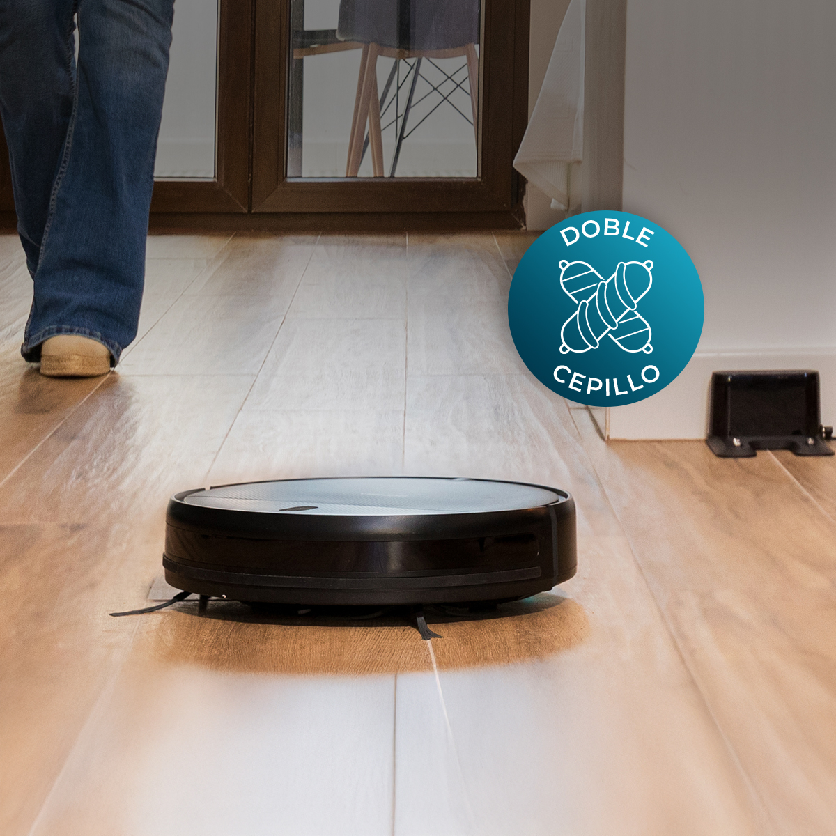 Un aspirapolvere robot nero su pavimento in legno. Una persona cammina sullo sfondo. C'è un logo con 'DOBLE CEPILLO' a destra.