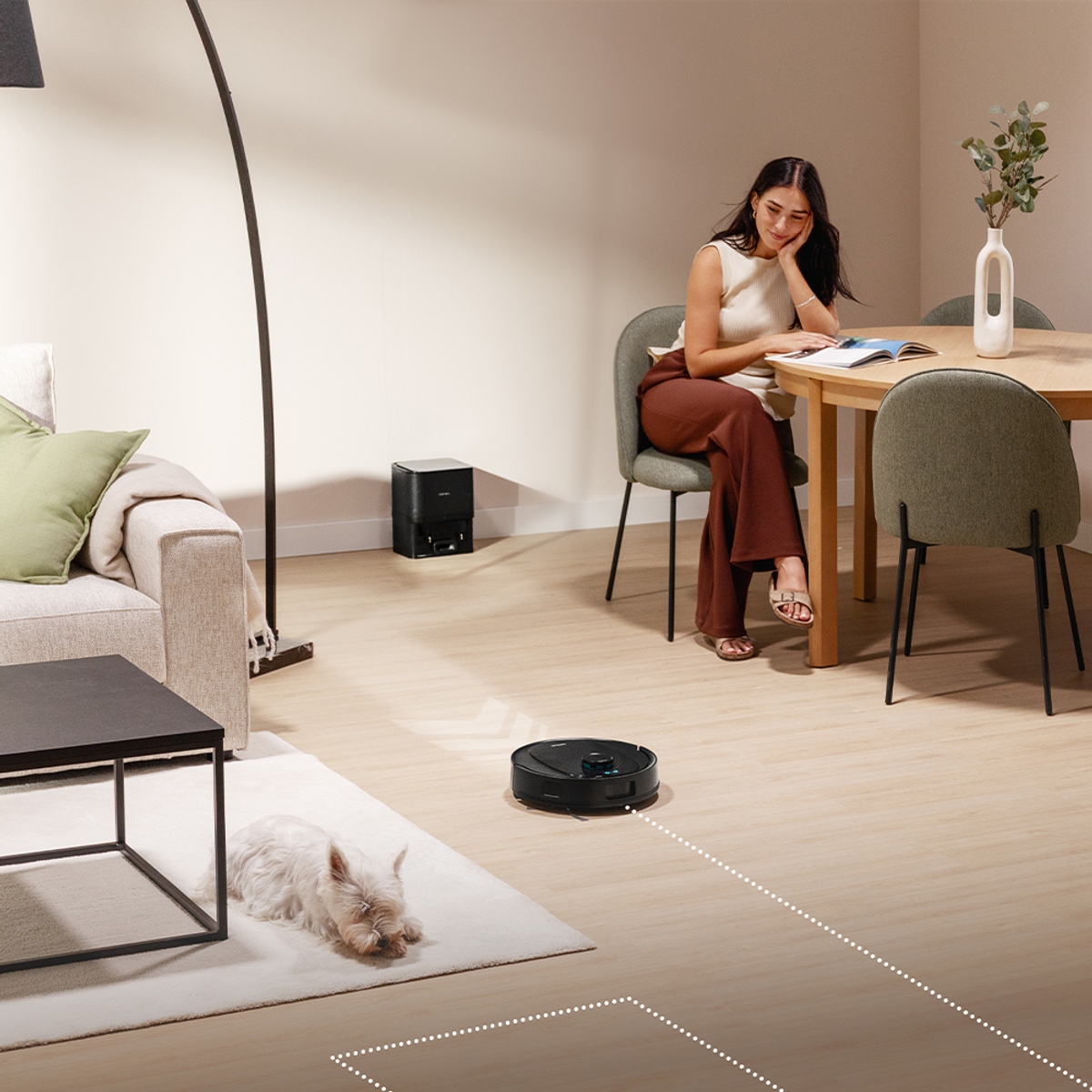 Una donna legge a tavola. Un aspirapolvere robot pulisce il pavimento. Un cane è su un tappeto. Una stazione di ricarica è dietro.