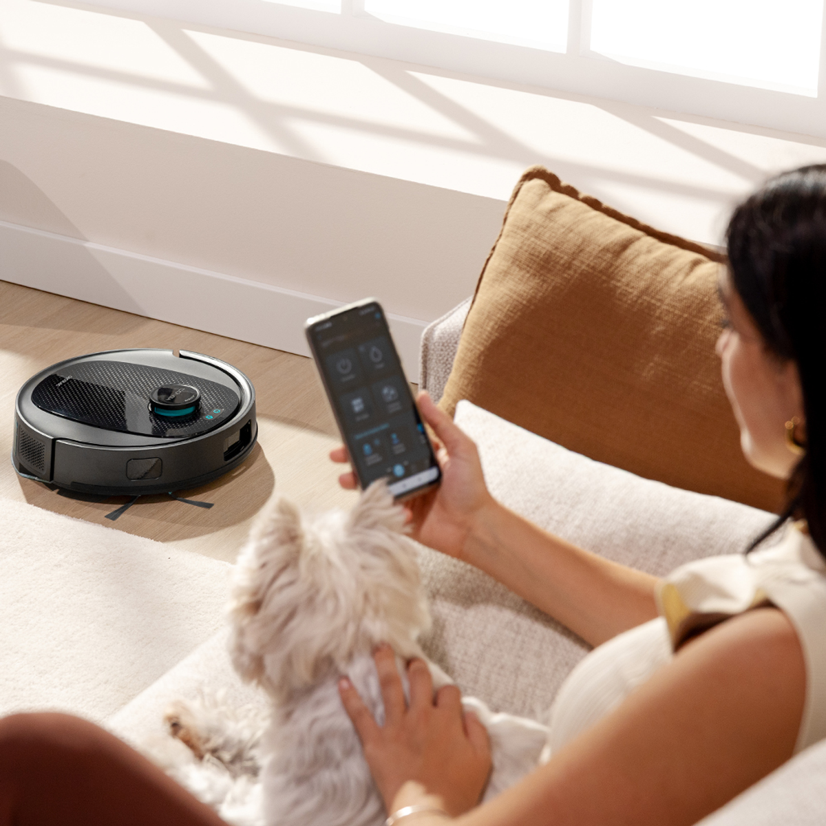 Una donna sul divano usa un telefono per controllare un aspirapolvere robot. Un cane bianco è sul divano. Una finestra sullo sfondo.