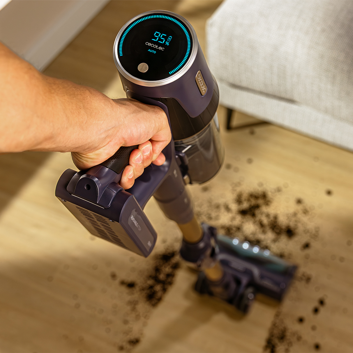 Una persona tiene un aspirapolvere senza fili su un pavimento in legno con sporco. Il display mostra la percentuale di batteria.