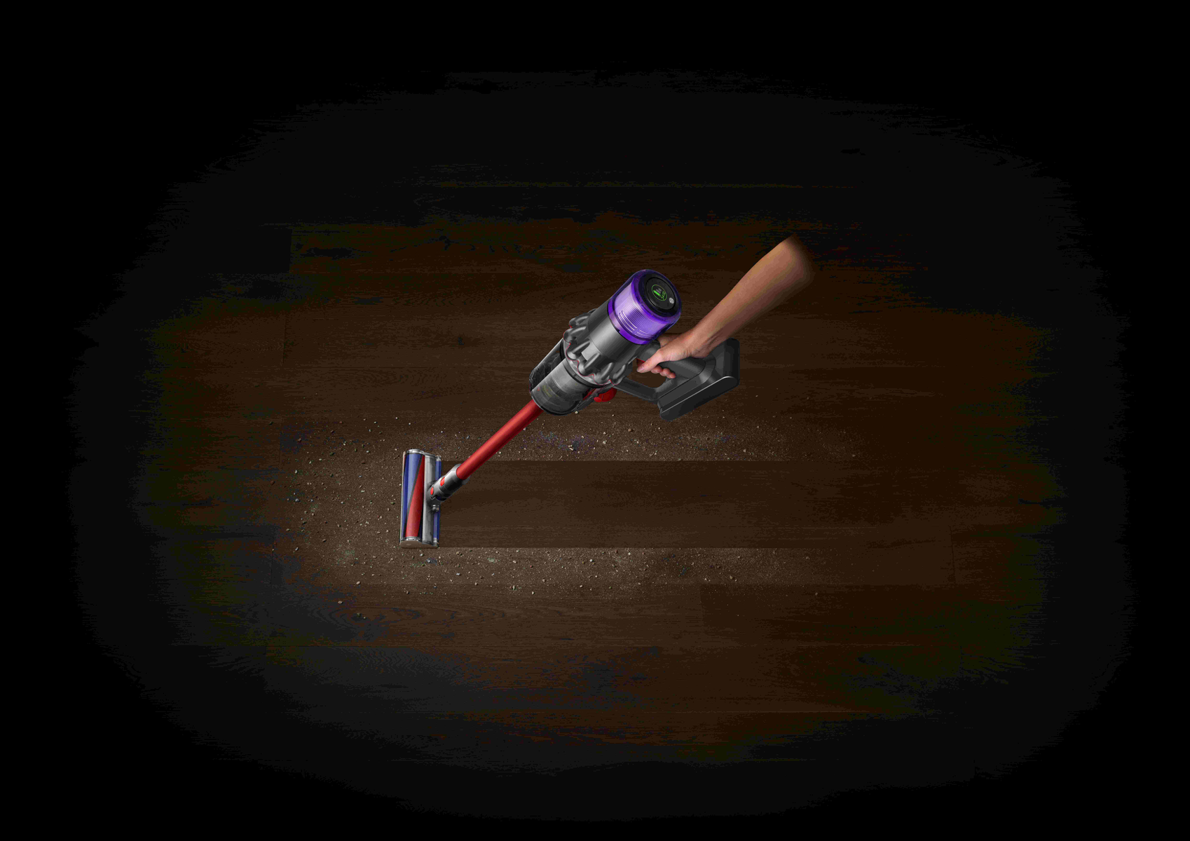 Eine Hand hält einen roten und silbernen kabellosen Staubsauger über einem Holzboden mit Staub. Der Staubsauger hat eine violette Oberseite.